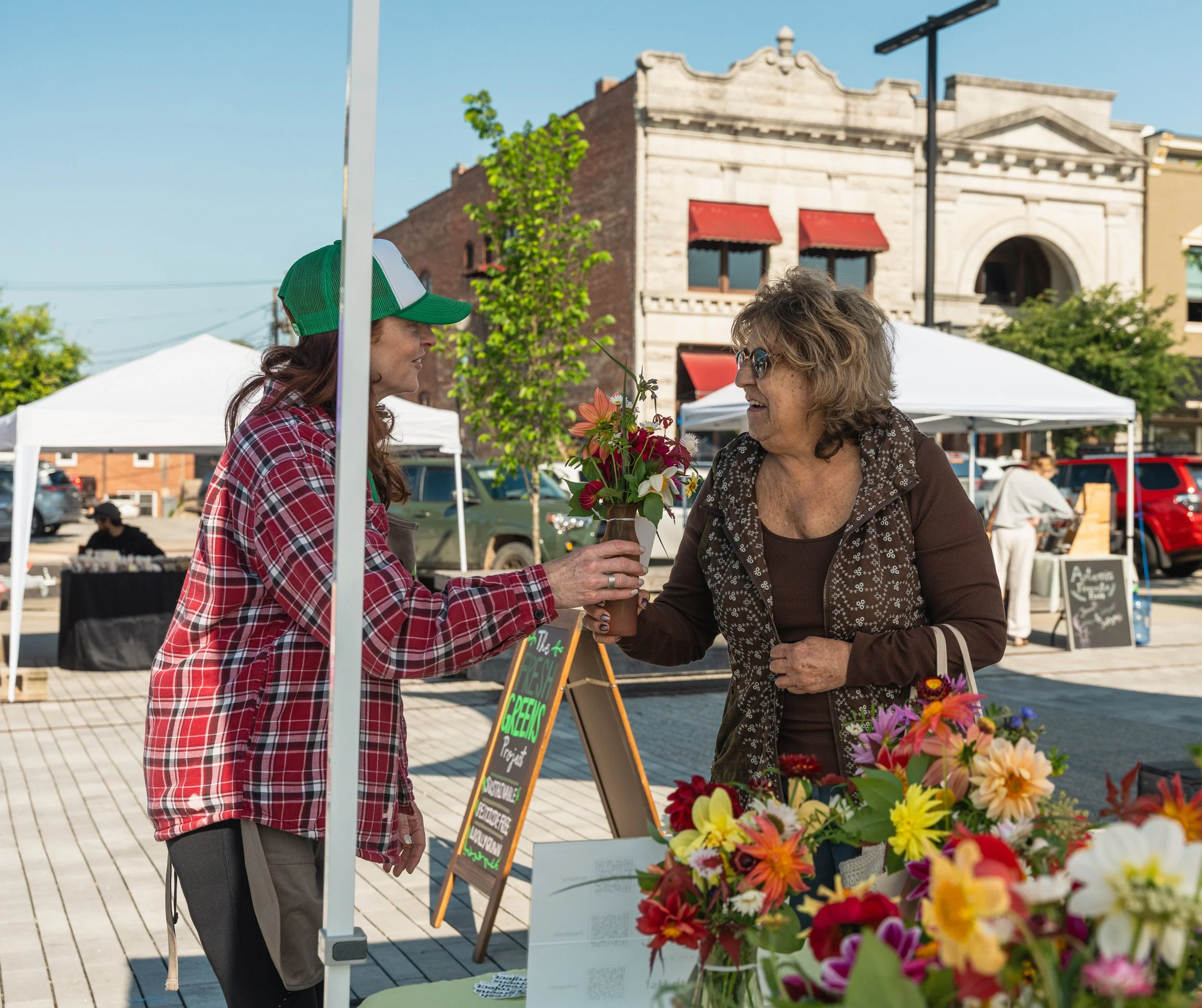 Two women exchanging flowers at an outdoor market with tents and historic brick buildings in the background.