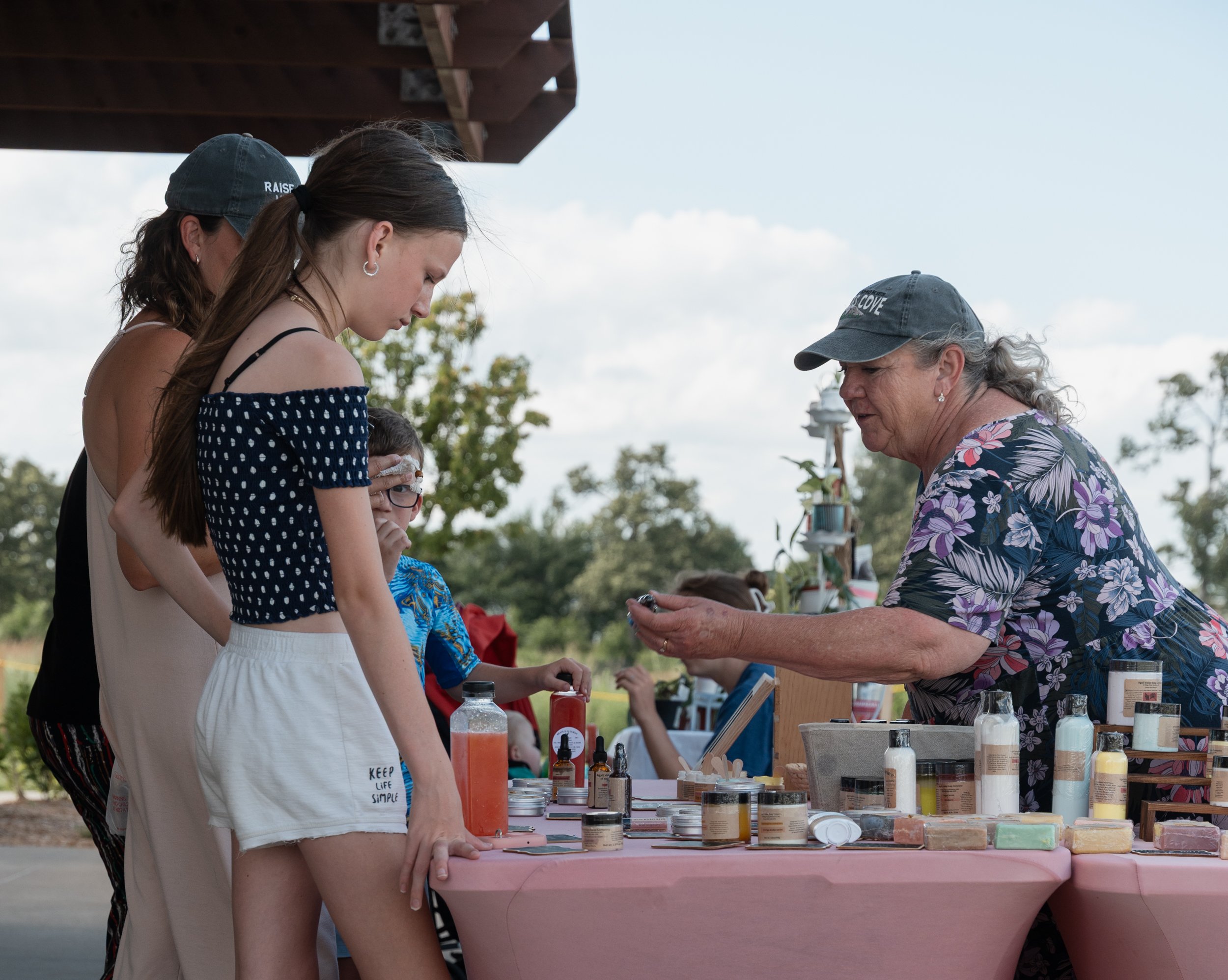 Several people gathered at an outdoor market stall with various bottles and containers of colorful powders and liquids, possibly for DIY or craft projects.