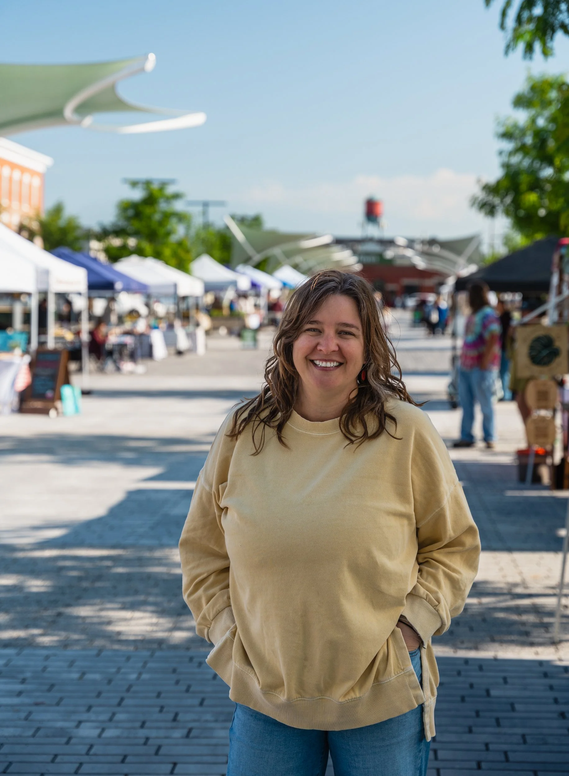A woman with long wavy brown hair and a beige sweatshirt standing outdoors at a market on a sunny day, smiling with a row of tents and people in the background.