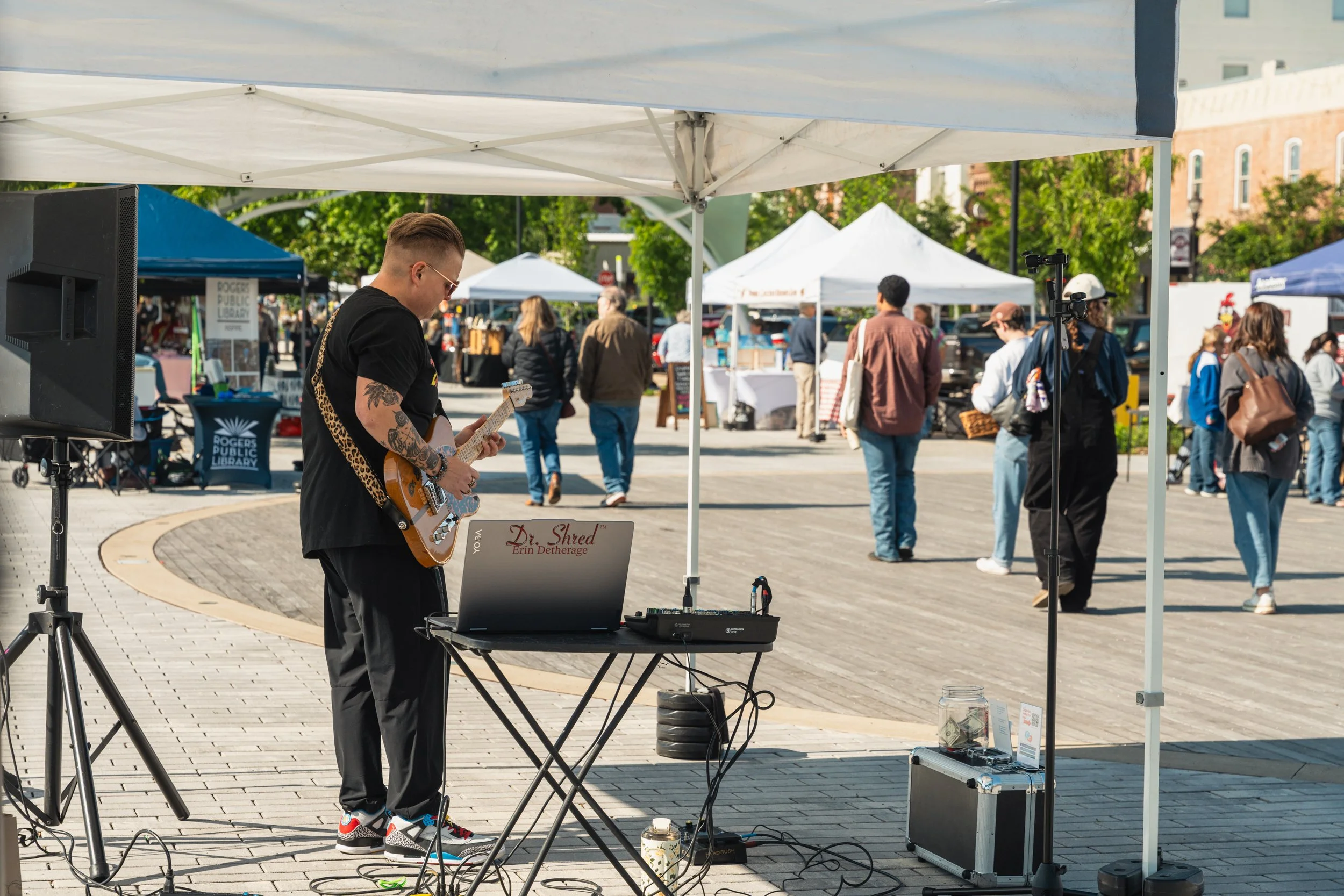 A musician with tattoos and sunglasses playing an electric guitar under a white canopy at an outdoor event, with people walking and browsing booths in the background.