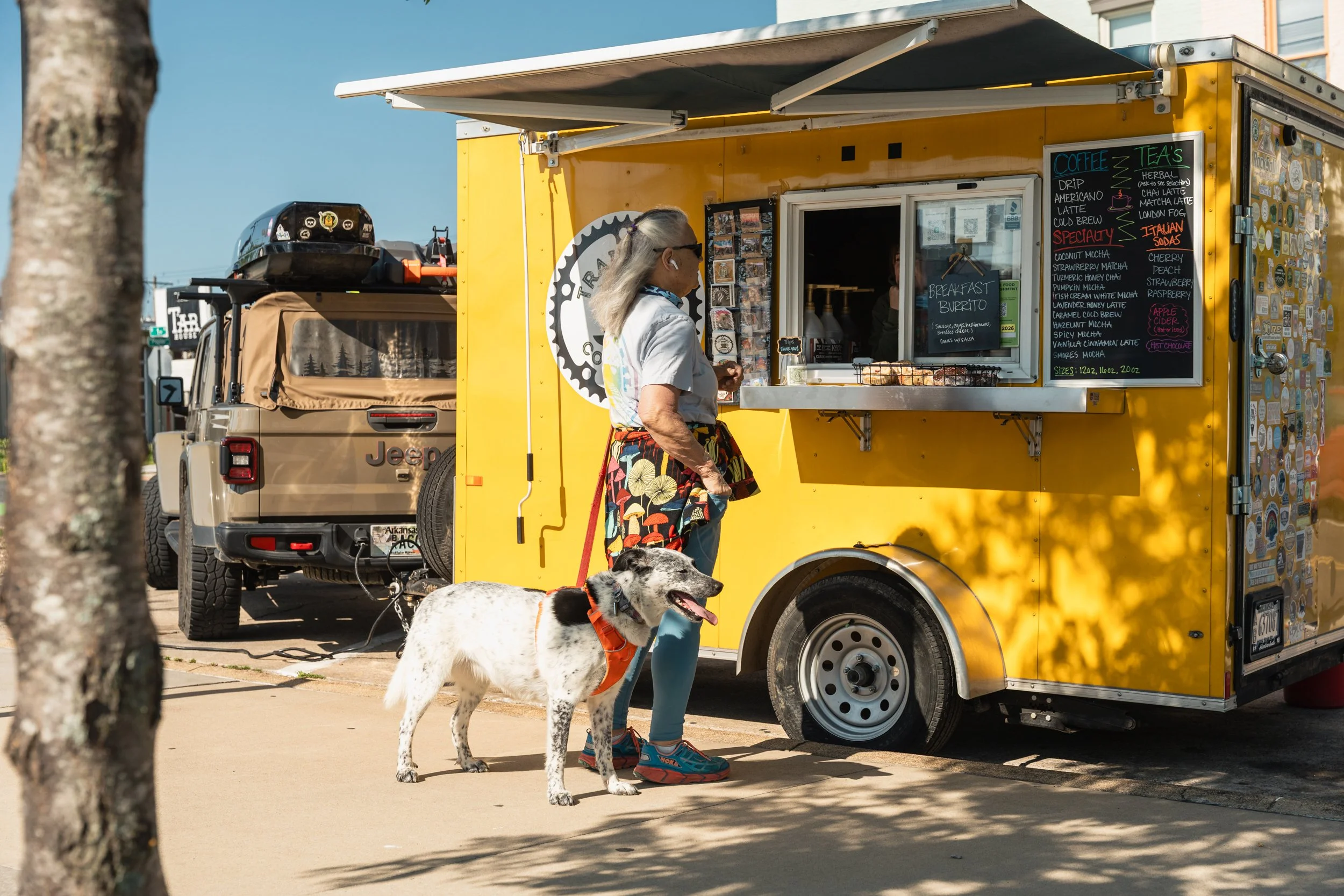 A woman with glasses and gray hair, carrying a bag, stands at a yellow food truck with a menu board. She has a dog on a leash with black and white fur and orange harness. The food truck offers coffee, teas, specialty drinks, and breakfast items, with decorative stickers on the side. Part of a brown vehicle is visible behind the food truck.