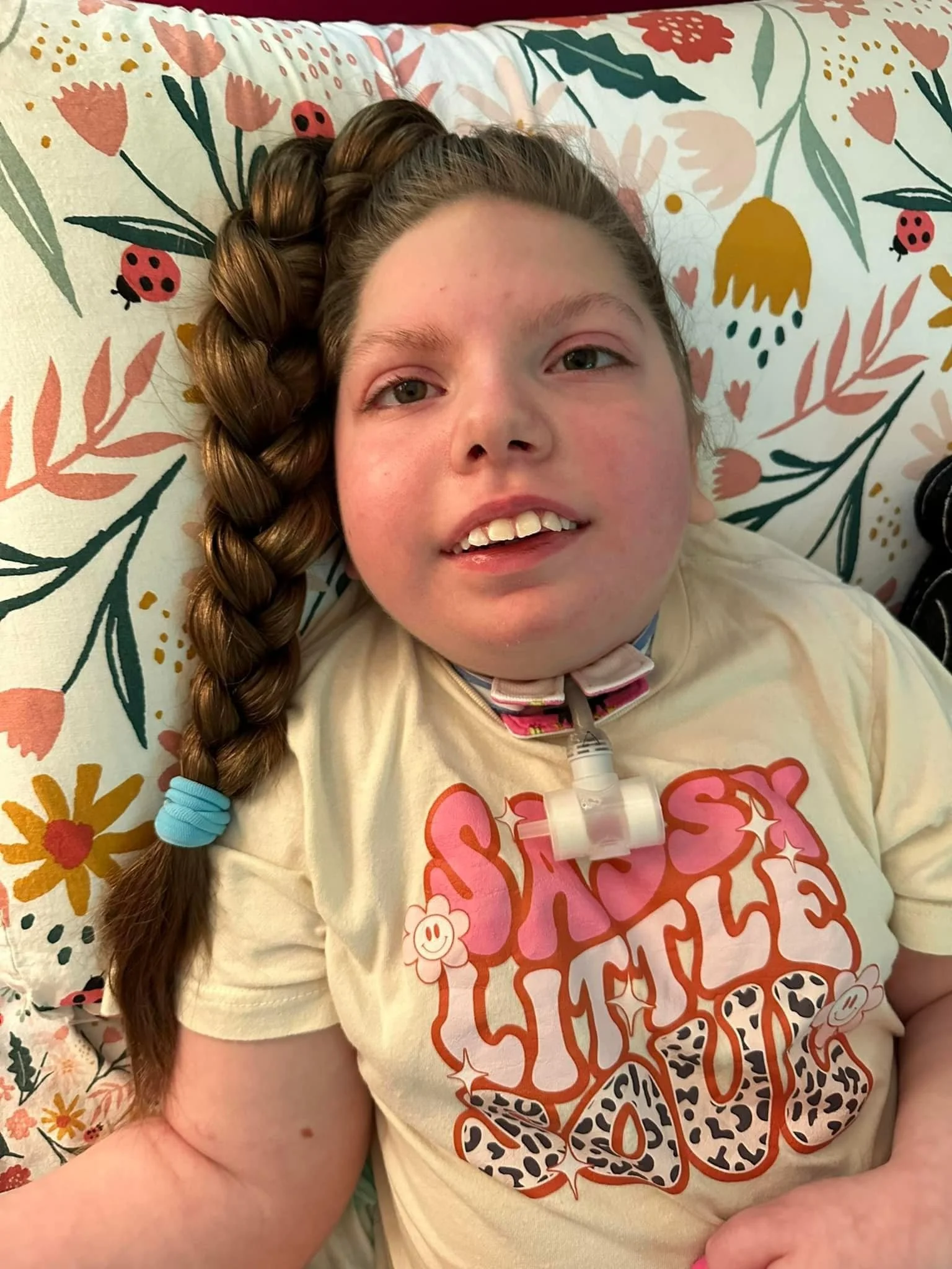 A girl with long braided hair lying in bed with a colorful floral and ladybug patterned bedsheet. She has a medical device attached, and is wearing a yellow T-shirt with orange and leopard print lettering.