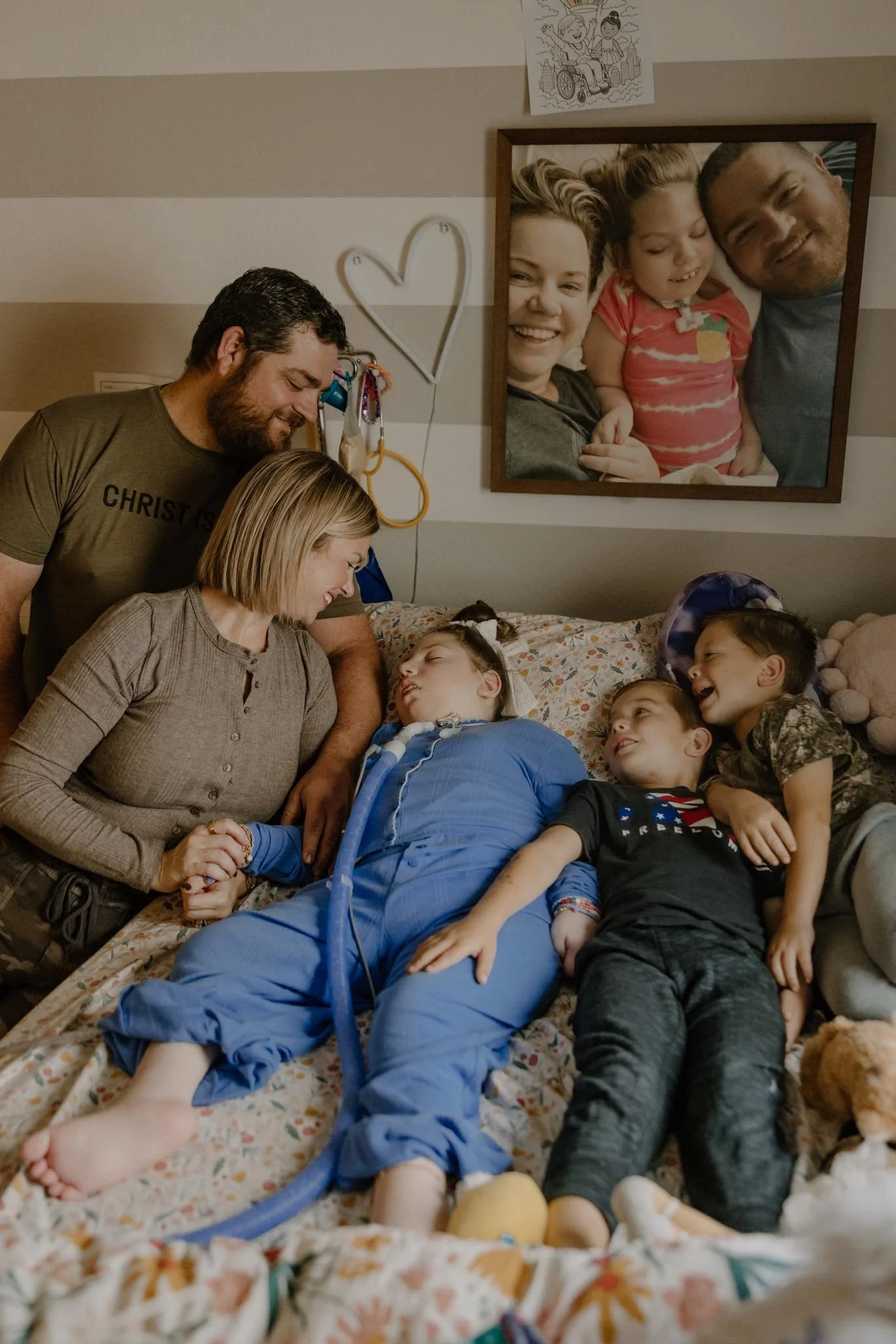 A family gathered in a hospital room. They are smiling and comforting a young girl who appears to be sleeping. A large framed photo of the family is on the wall, along with children's drawings and hospital equipment.