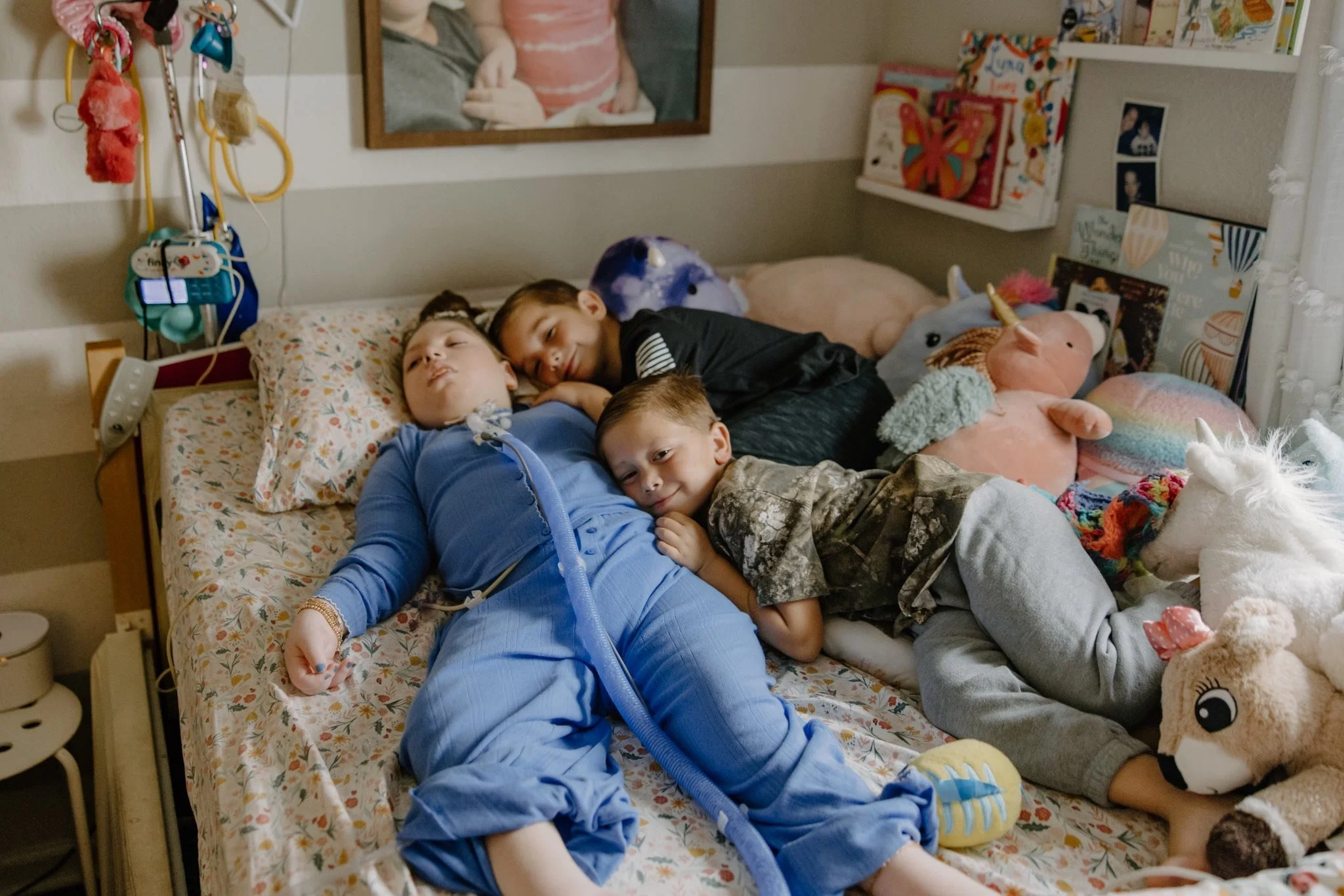 Three children in a hospital bed with plush toys and stuffed animals, resting and smiling.