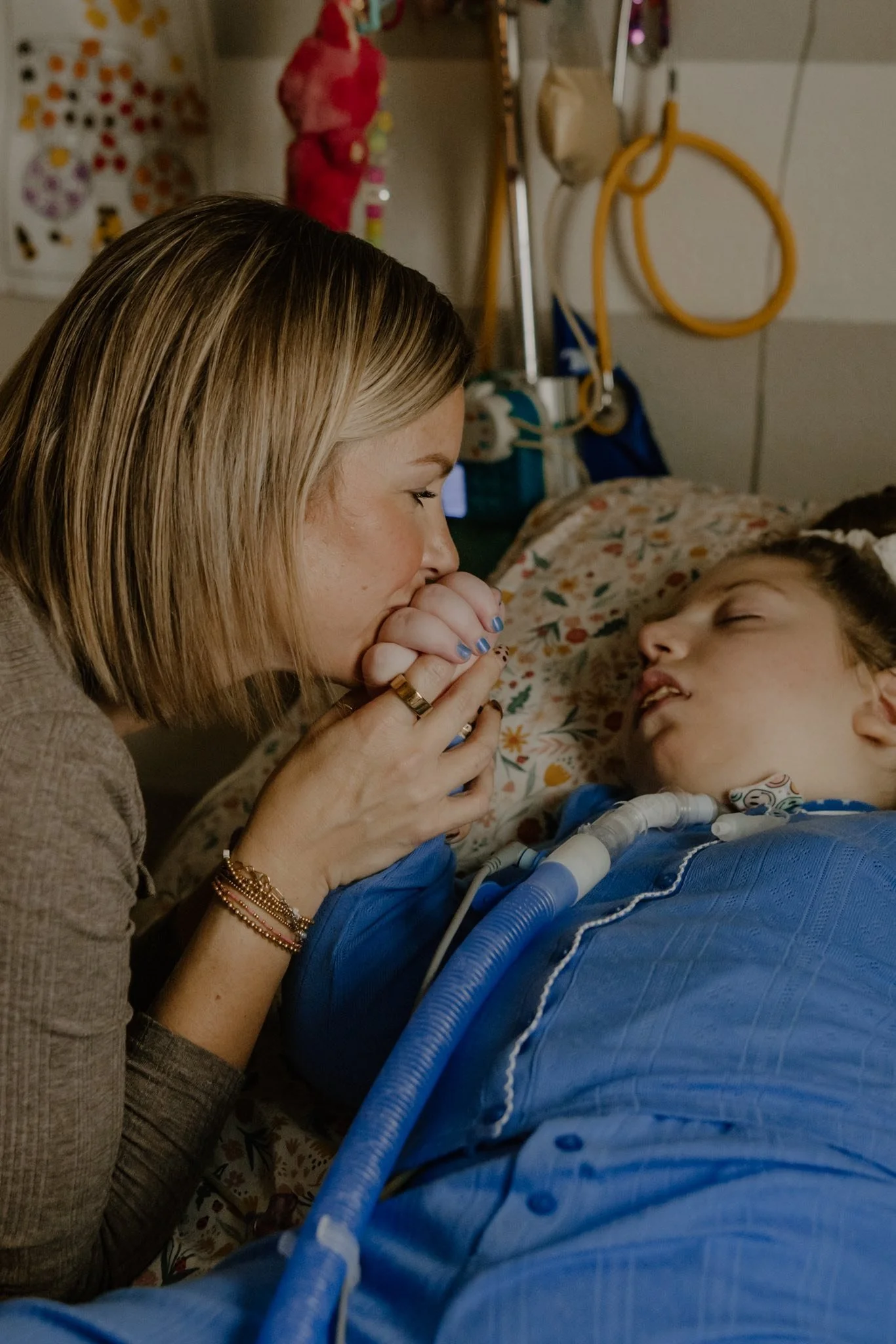A woman with blonde hair holding the hand of a young girl in a hospital bed. The girl appears to be sleeping or unconscious, with medical tubes attached. The background shows medical equipment and colorful wall decorations.