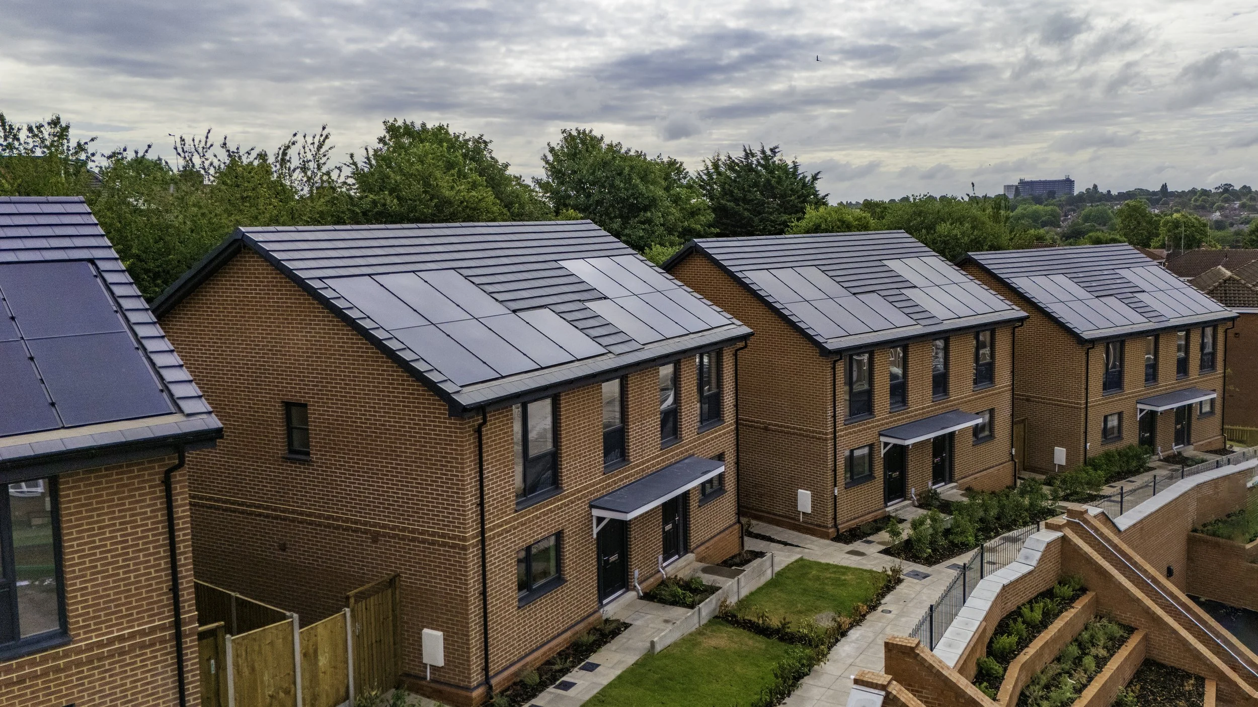 Multiple modern brick townhouses with dark-colored solar panels on their roofs, surrounded by green trees and a cloudy sky in the background.