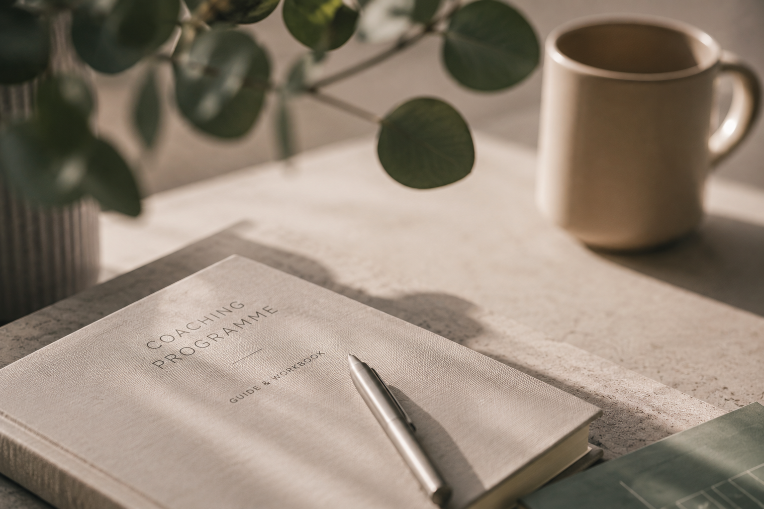A coaching guide and workbook with a silver pen resting on it, placed on a light-colored textured surface. There is a book or magazine partially visible beside it and a potted plant with green leaves casting shadows nearby, along with a cream-colored coffee mug.