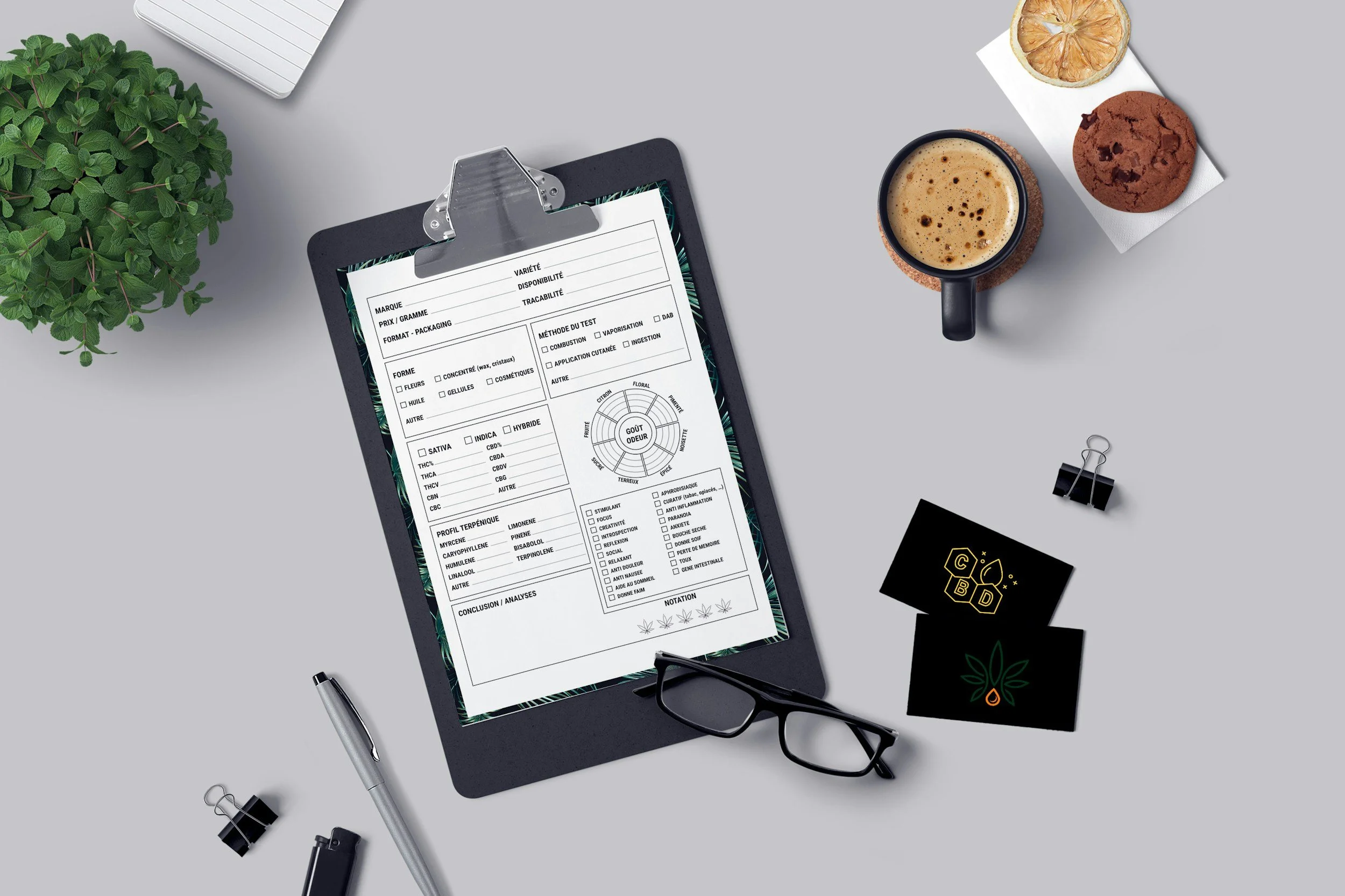 A workspace with a clipboard holding a form or checklist, a pair of eyeglasses, a black CBD product box, a silver pen, and scattered binder clips. There is a small potted plant, a notepad, cups of coffee, and snacks including dried lemon slices and chocolate chip cookies on the light gray surface.