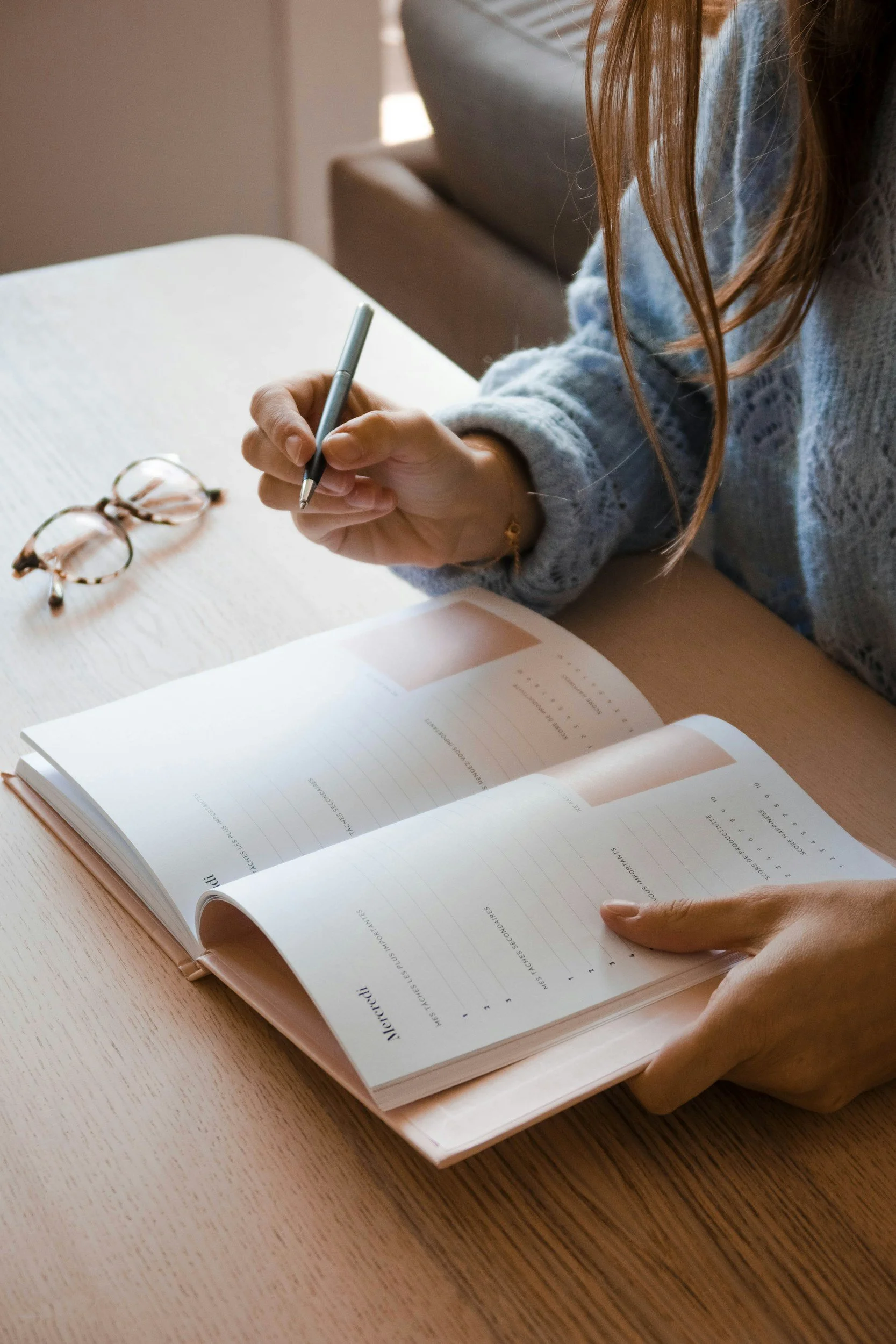A workspace with a clipboard holding a form or checklist, a pair of eyeglasses, a black CBD product box, a silver pen, and scattered binder clips. There is a small potted plant, a notepad, cups of coffee, and snacks including dried lemon slices and chocolate chip cookies on the light gray surface.