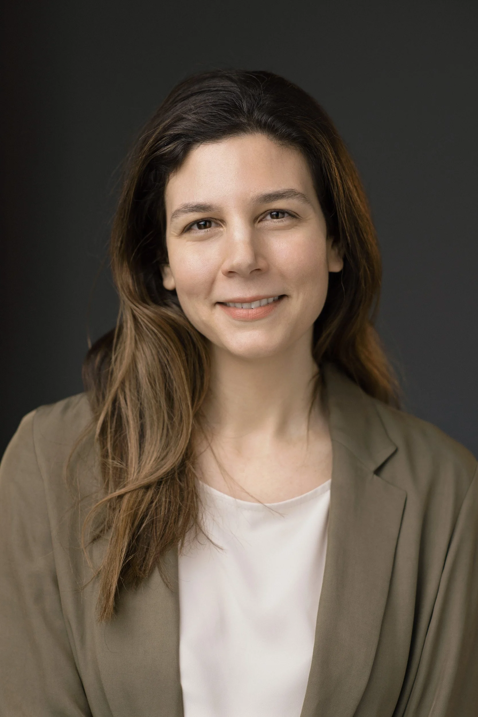 Portrait of a woman with long wavy brown hair, smiling, wearing a beige blazer and a white shirt, against a dark background.