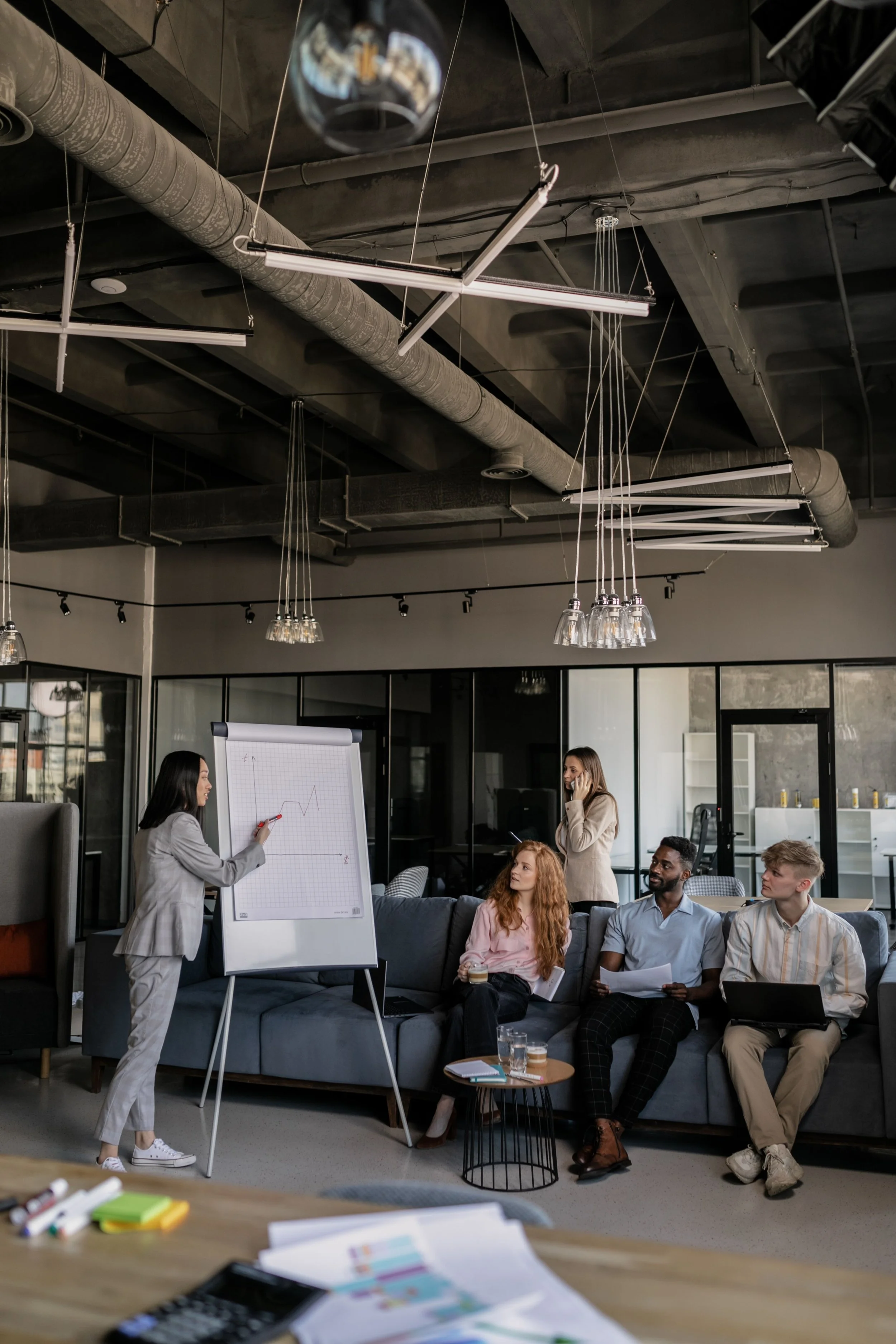 A group of five diverse professionals in a modern office, participating in a presentation. One woman is standing at a whiteboard with a graph, pointing to it with a marker. Four others, three women and one man, are seated on a couch, engaged and listening, with some holding papers and a laptop. The office has an industrial ceiling with exposed ducts and hanging lights, and large windows.