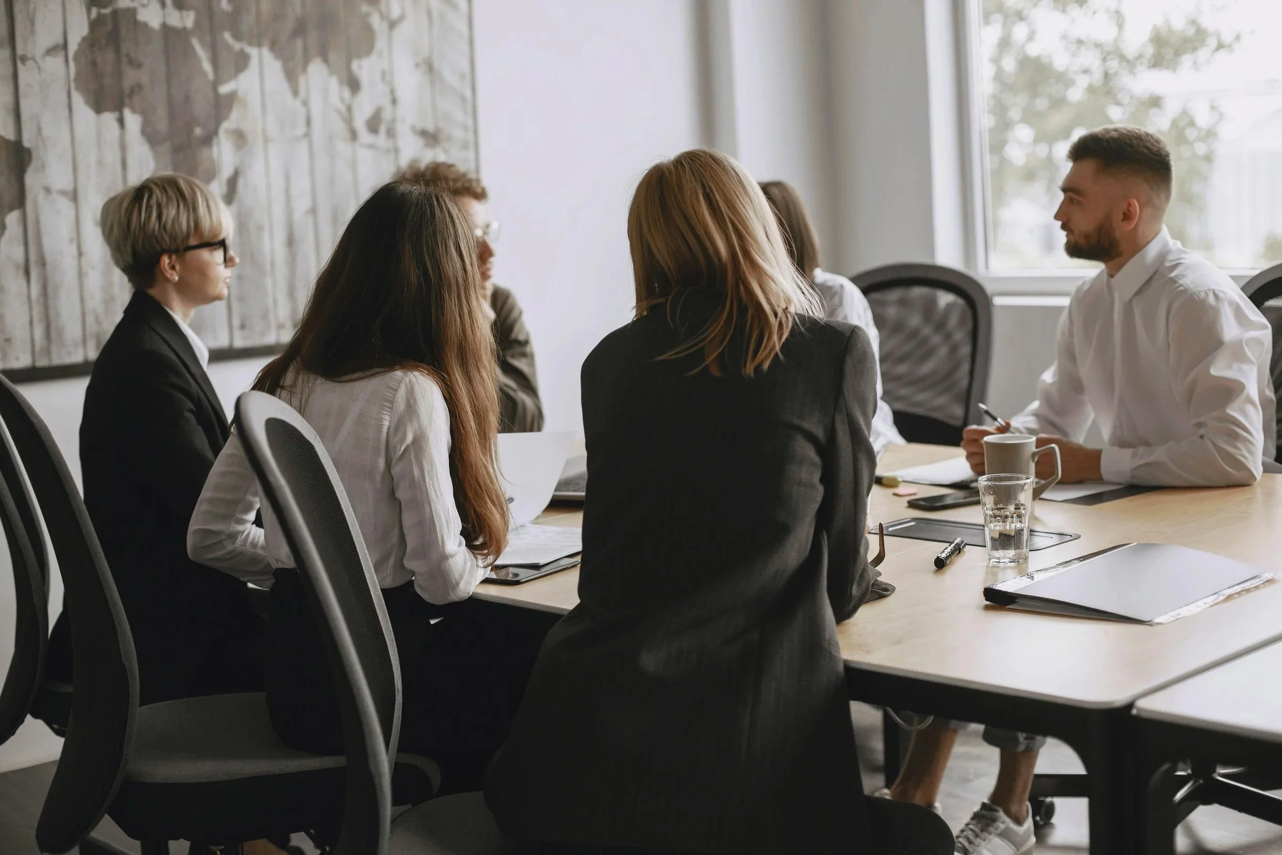 Group of six people sitting around a conference table in a meeting room with large windows, engaged in discussion.