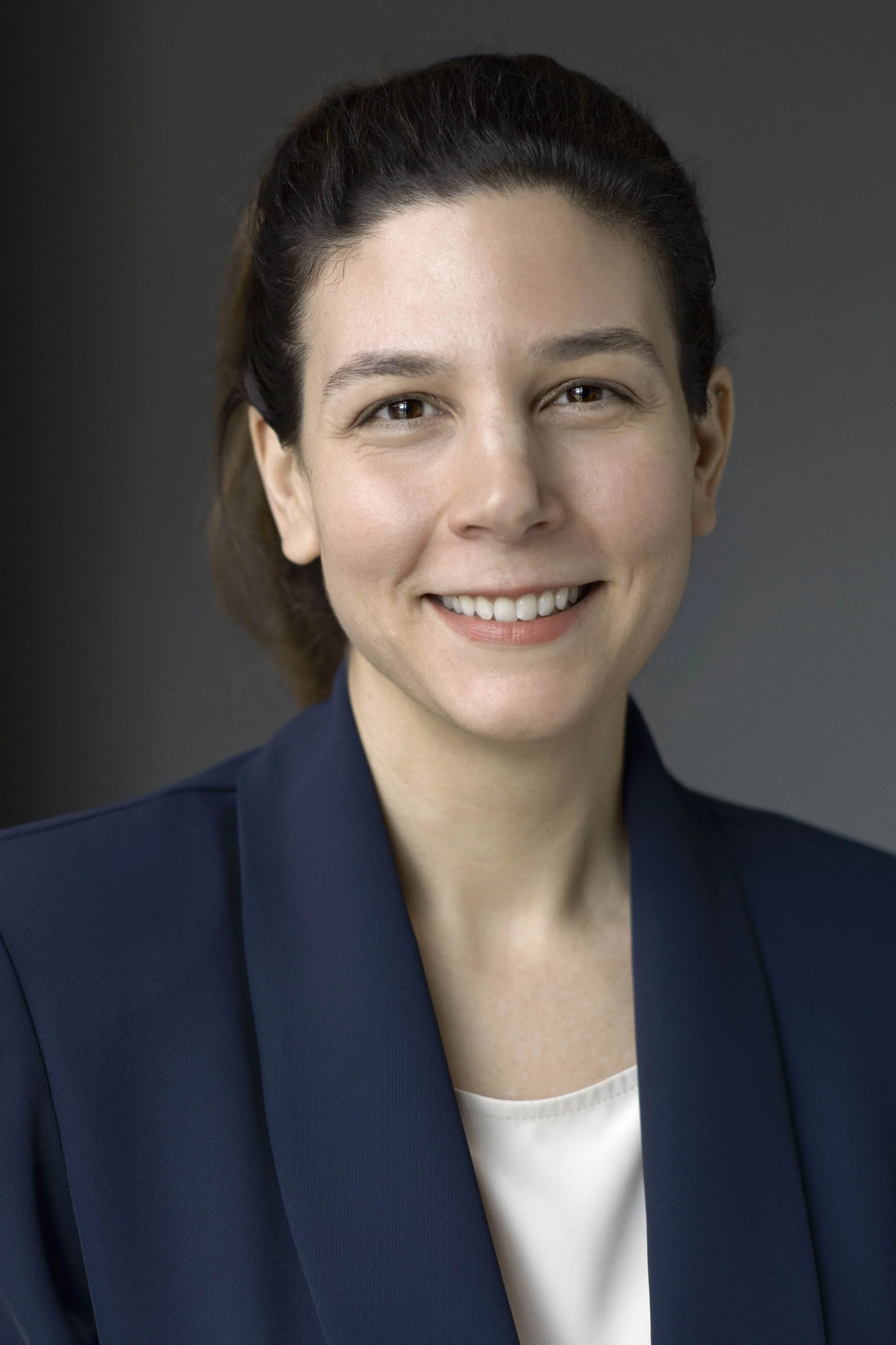 Close-up portrait of a smiling woman with dark brown hair styled back, wearing a navy blazer and a white top, against a dark gray background.
