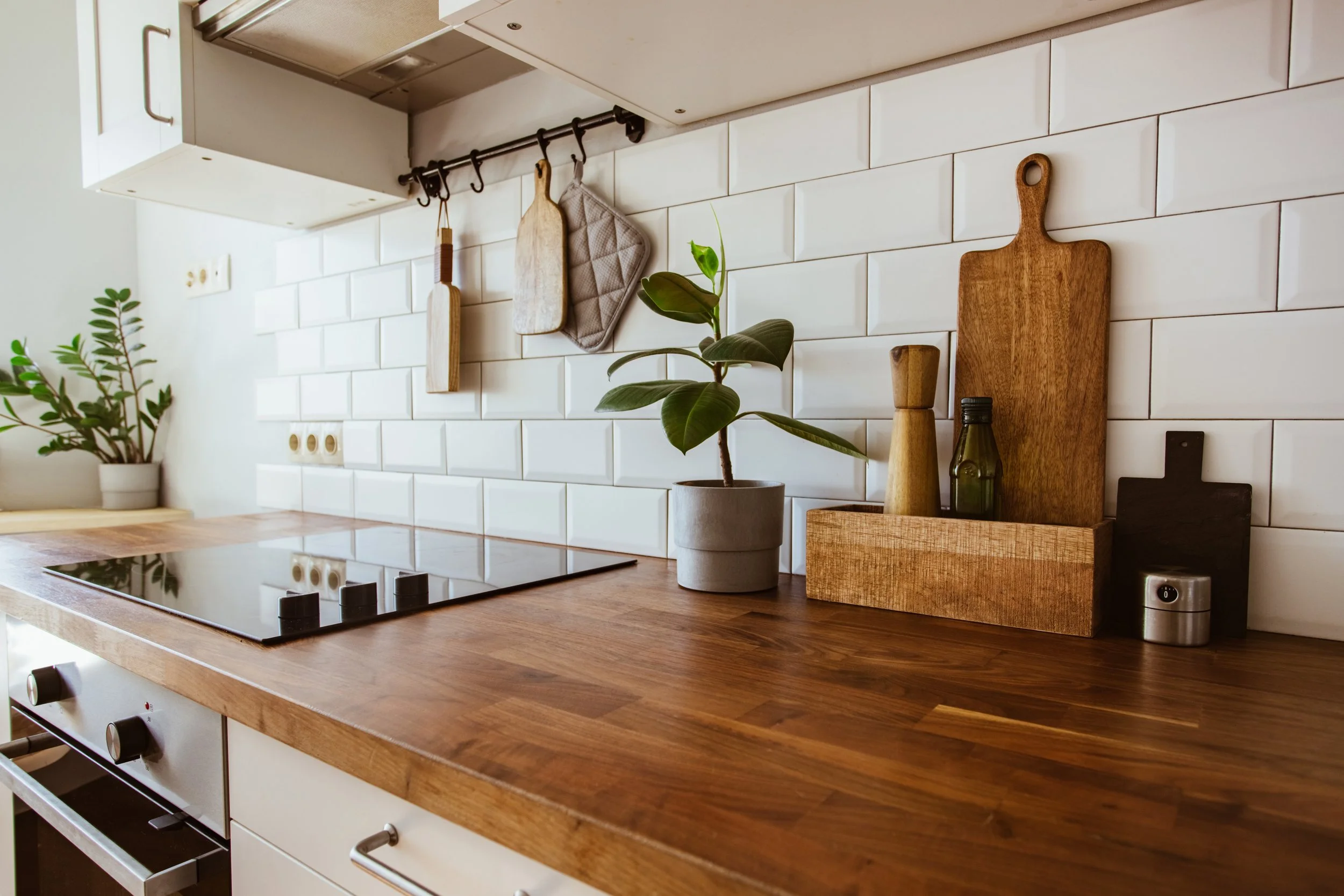A modern kitchen counter with a wooden surface, white tiled backsplash, potted plant, wooden cutting boards, a glass bottle, and various kitchen tools hanging on a rail.