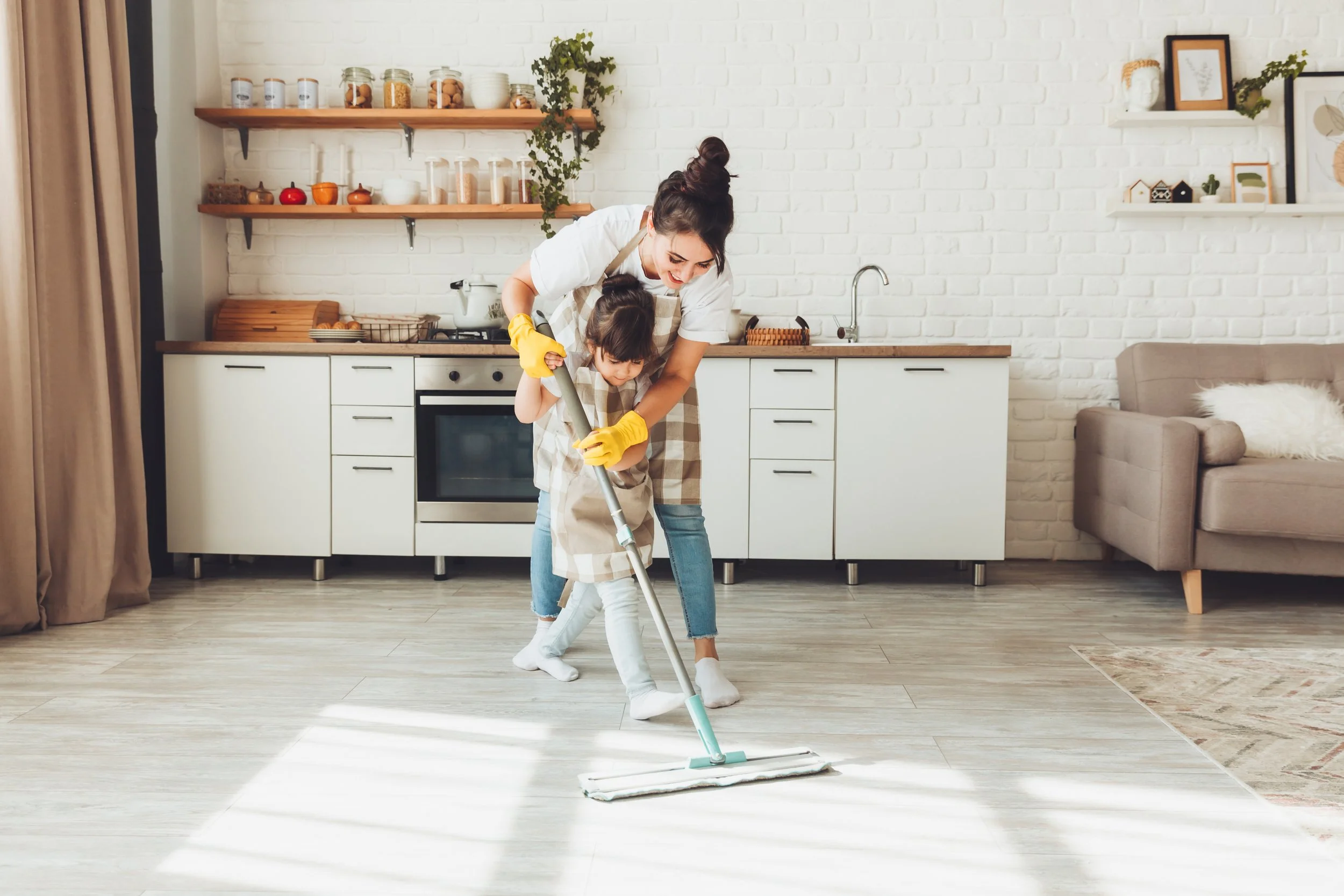 A woman and a young girl holding a mop together in a modern kitchen, cleaning the floor.