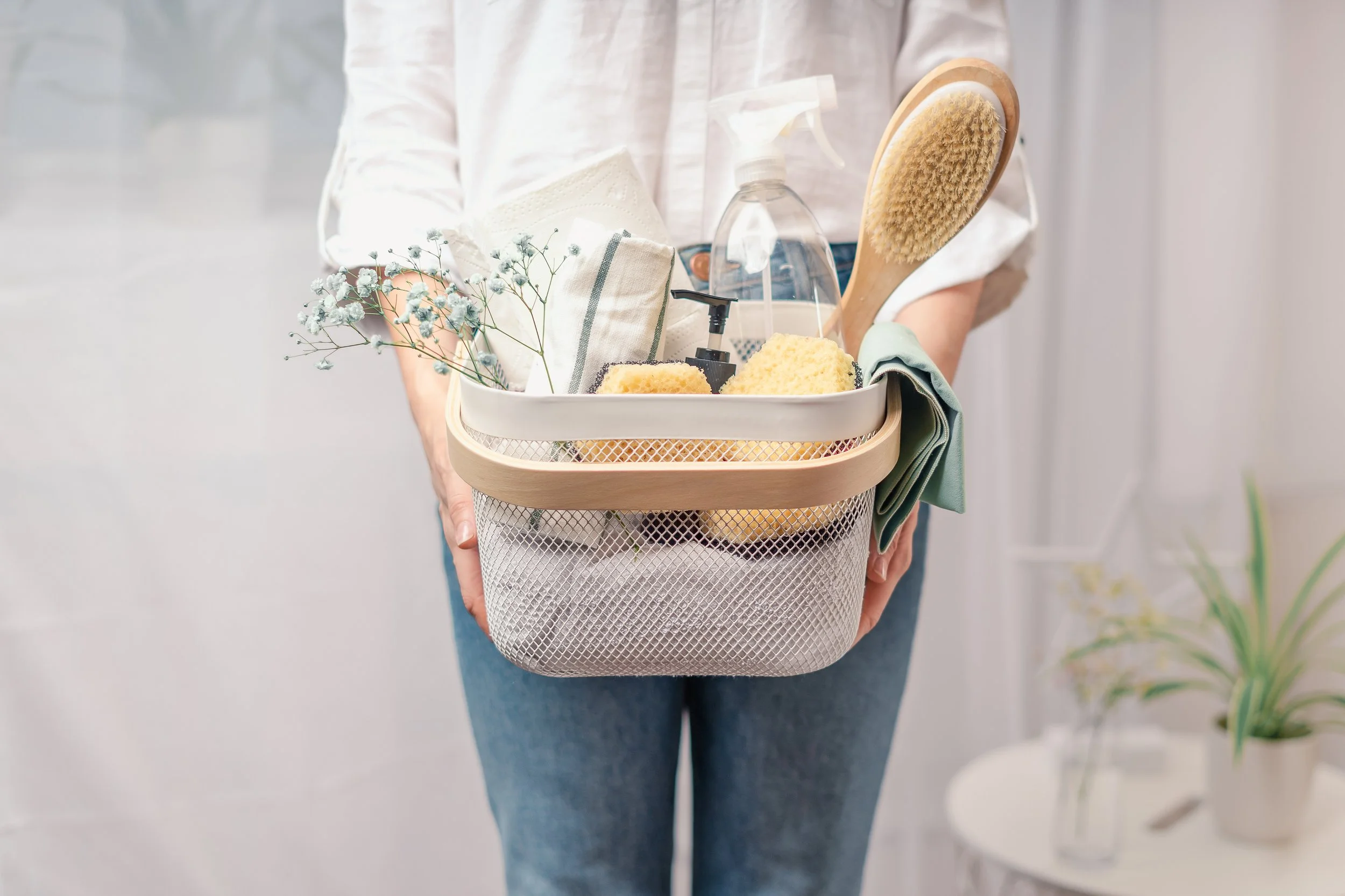 Person holding a cleaning supply caddy with a brush, spray bottle, cleaning sponges, flowers, and a towel.