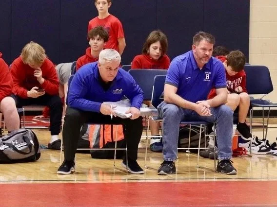 A group of young athletes and two coaches sitting on chairs along a gym wall during a sports event.