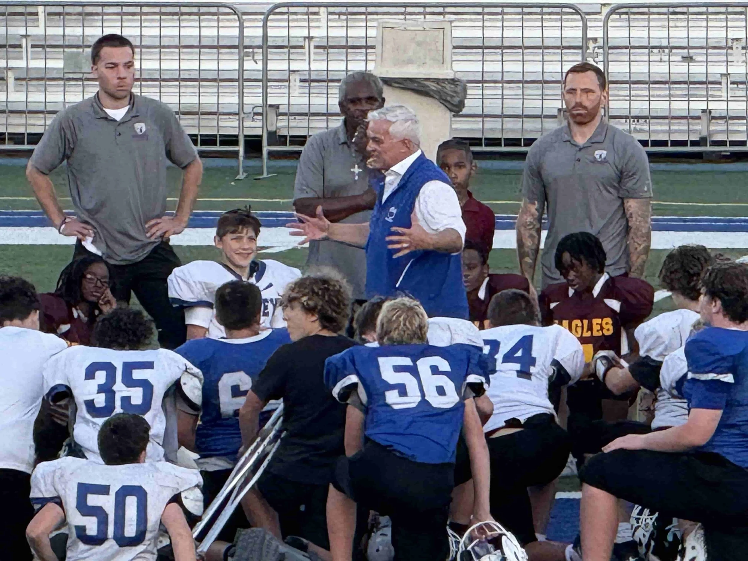 Bexley Middle School Football Team sideline of a field, with two men standing and talking in front. The men are dressed in gray shirts, and the players are in white and blue uniforms with numbers on the back.