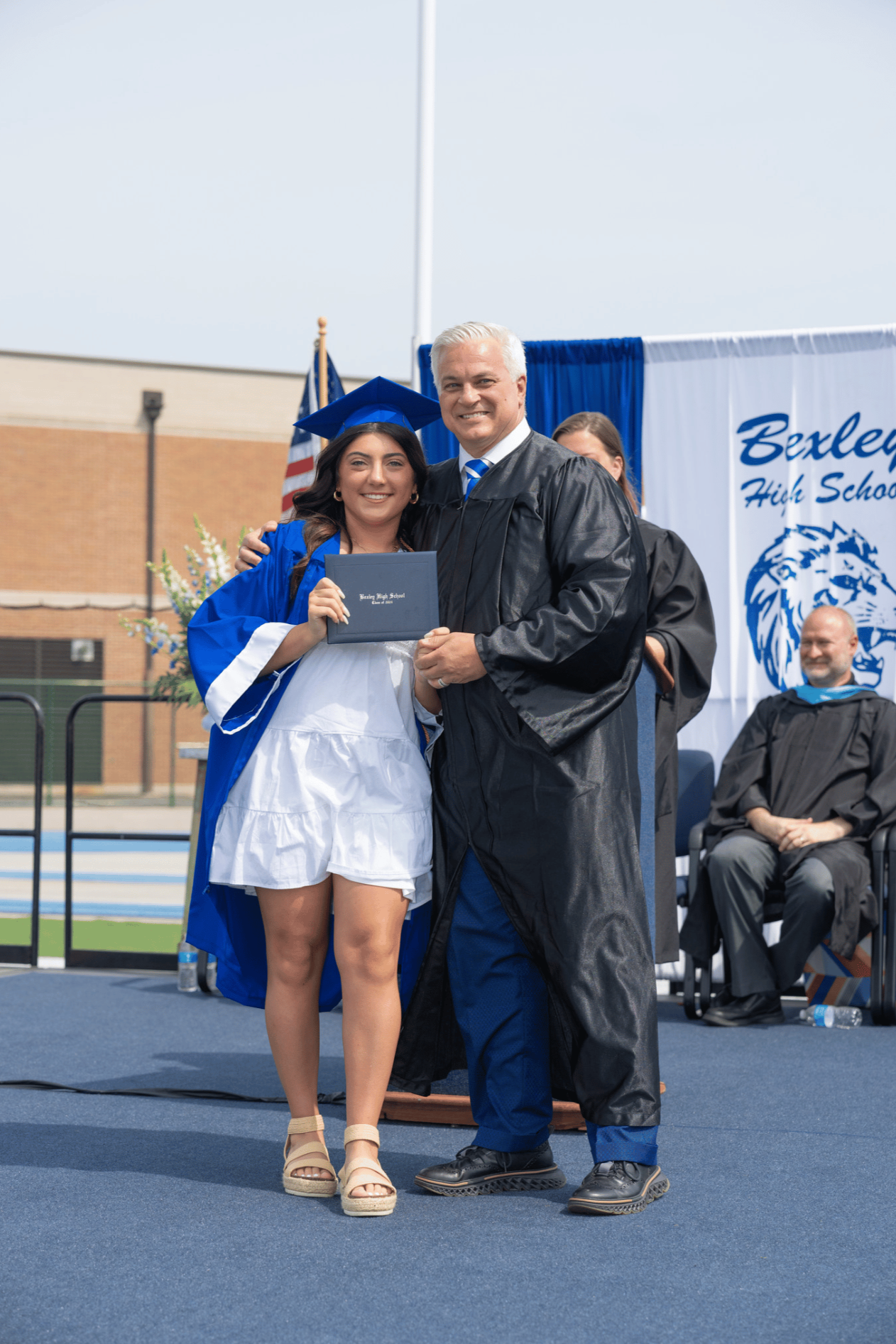 A young woman in a white and blue graduation gown and cap holding a diploma standing next to a man in a black academic gown at a high school graduation ceremony, with other people and a blue and white banner in the background.