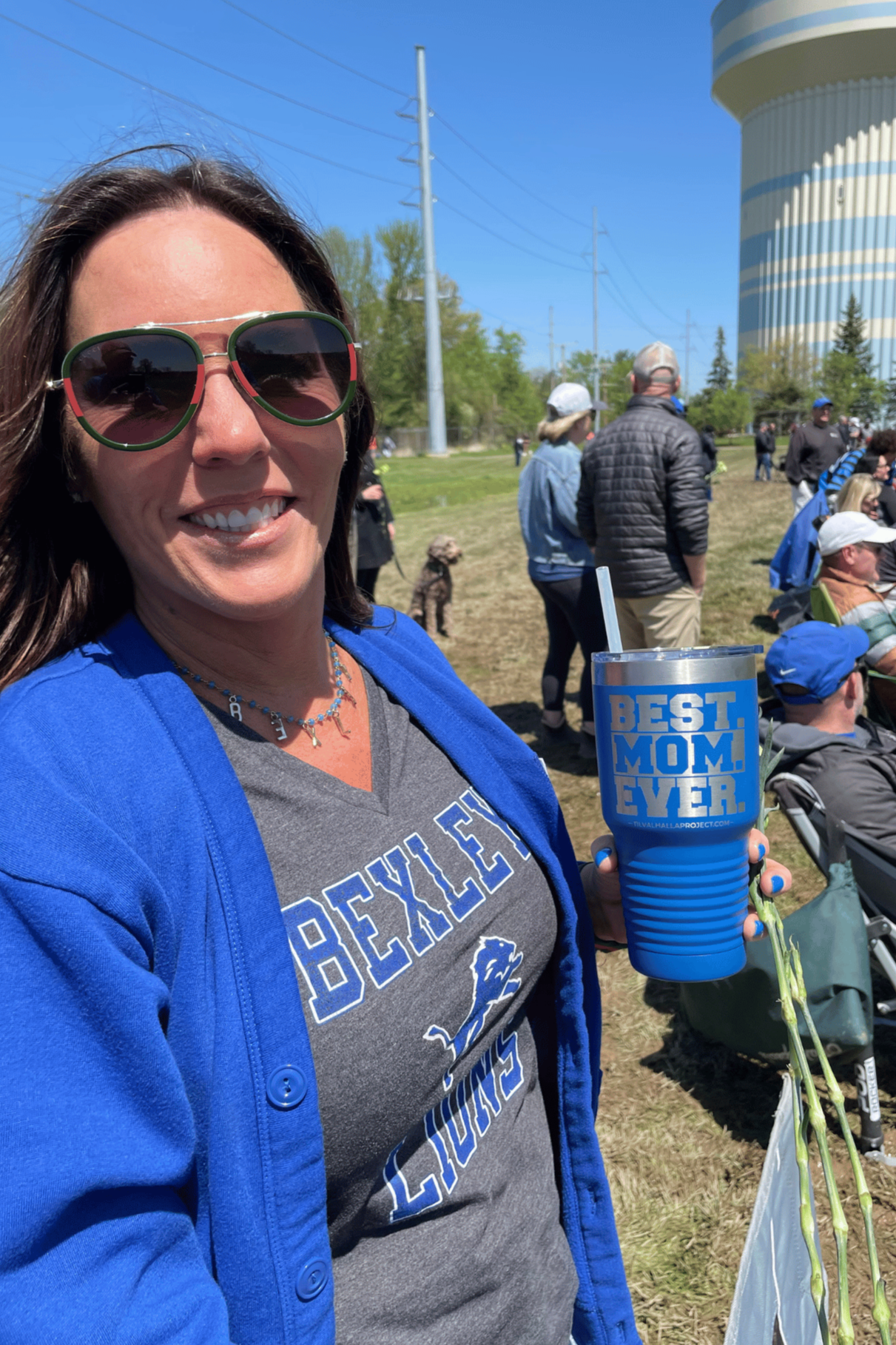 Smiling woman wearing sunglasses and a blue jacket, holding a blue tumbler with 'Best Mom Ever' written on it, at an outdoor event on a sunny day with people and a water tower in the background.