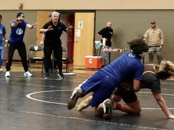 Bexley OH Middle School Ohio wrestlers competing on the mat, one in a blue uniform and the other in a black and yellow uniform, with spectators watching and reacting in the background.