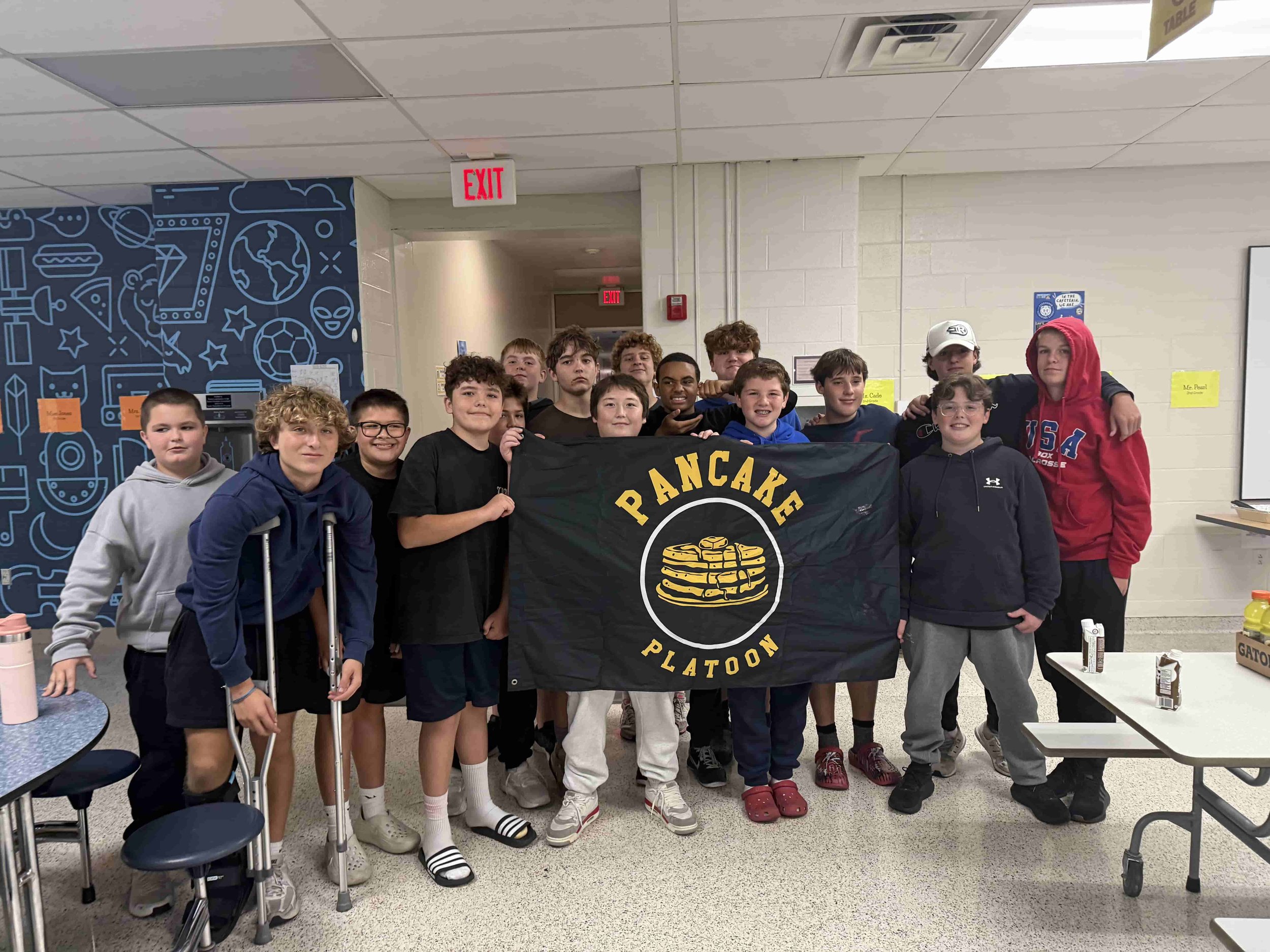 Bexley Middle School Football Team Community Service Bexley OH Ohio Bexley School Team in a cafeteria holding a black flag that reads 'Pancake Platoon,' with some smiling and others looking serious, some wearing glasses, hoodies, and sportswear.