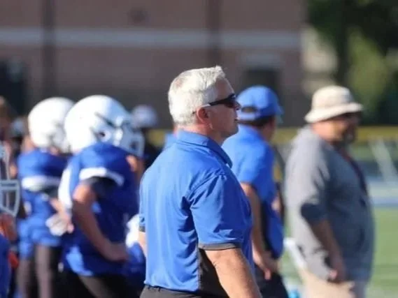 Bexley Ohio OH Middle School Football Coach or referee standing on a football field with players in blue uniforms and helmets in the background.