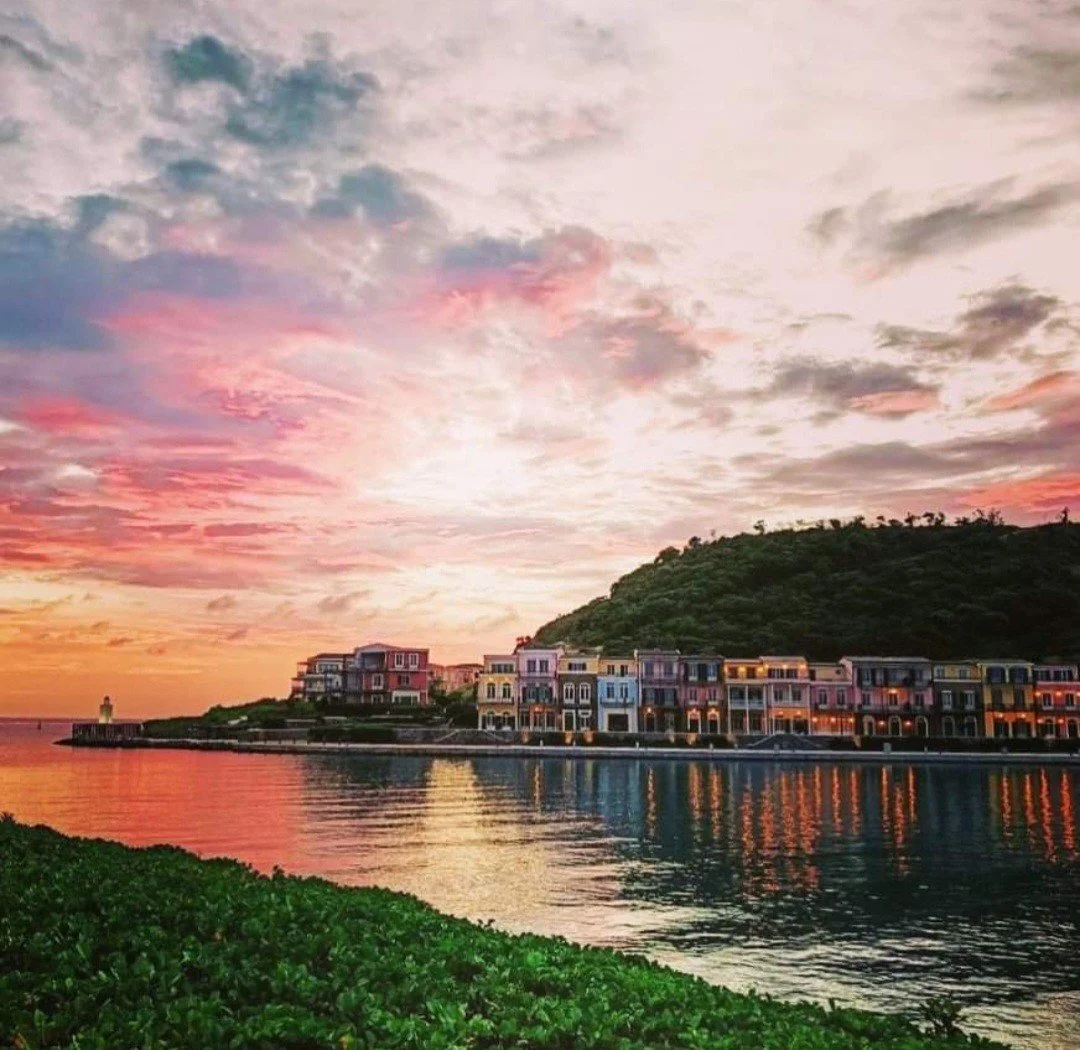 Colorful sunset over a coastal town with calm water reflecting the buildings, green foliage in the foreground, and a hill in the background.