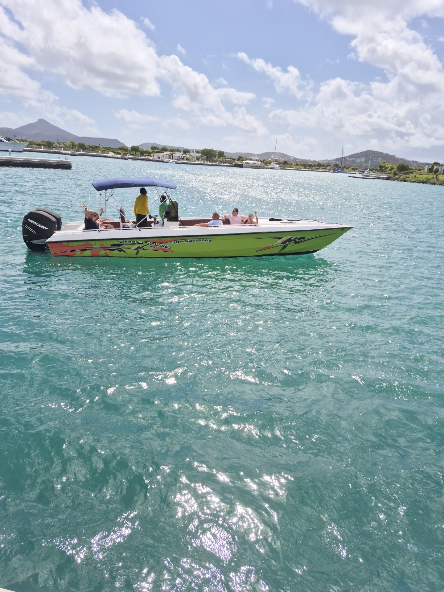 A boat with passengers on the water, with mountains and buildings in the background, under a partly cloudy sky.