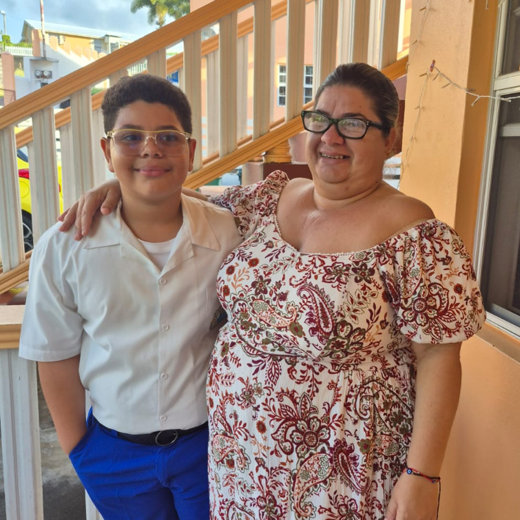 A boy and a woman standing together on a porch, smiling. The boy is wearing glasses, a white shirt, and blue pants, while the woman is wearing a floral-patterned dress and glasses.