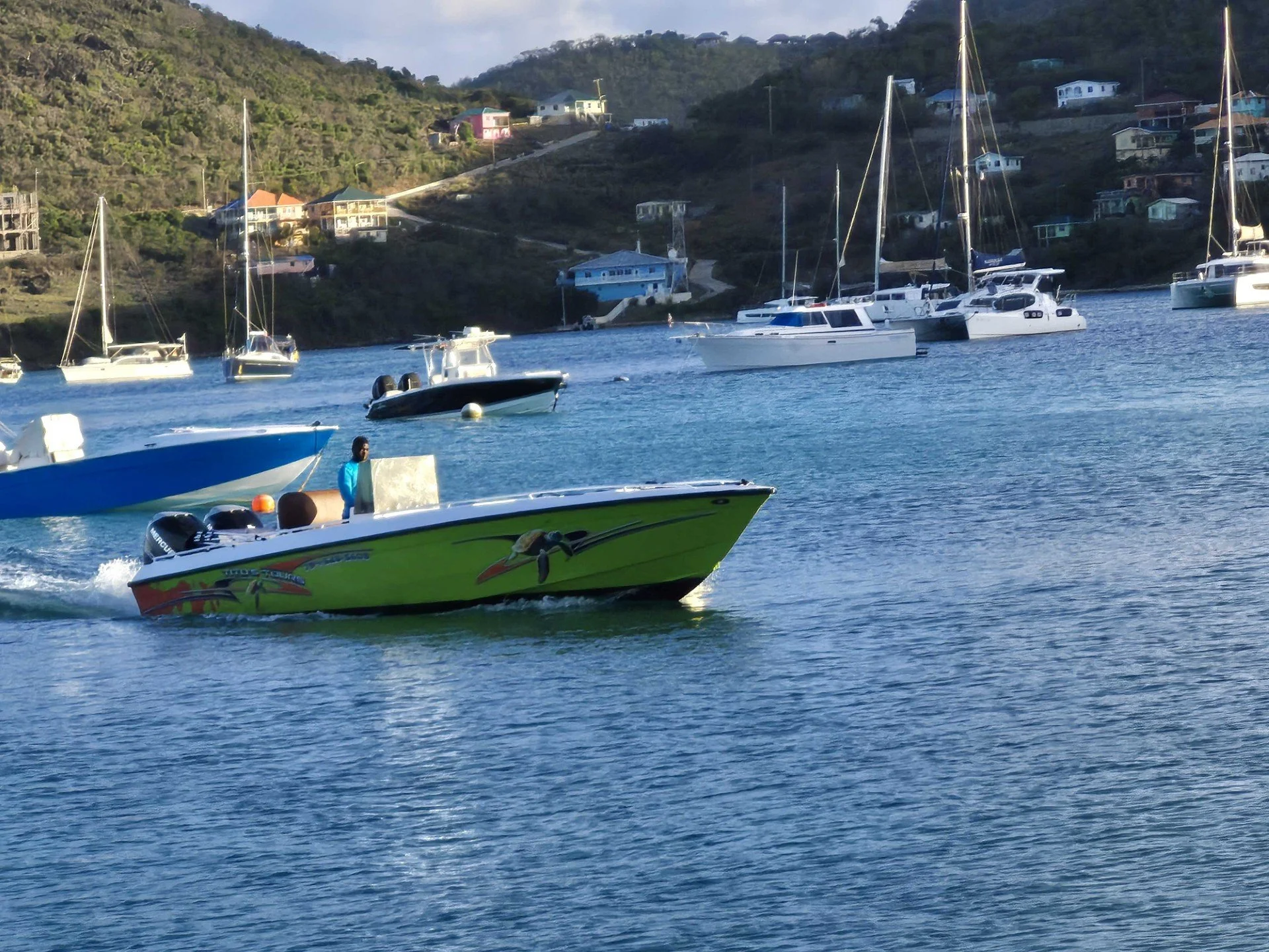 A motorboat with colorful decor speeds through the water with a person on board, surrounded by sailboats and yachts anchored near a hillside with houses.
