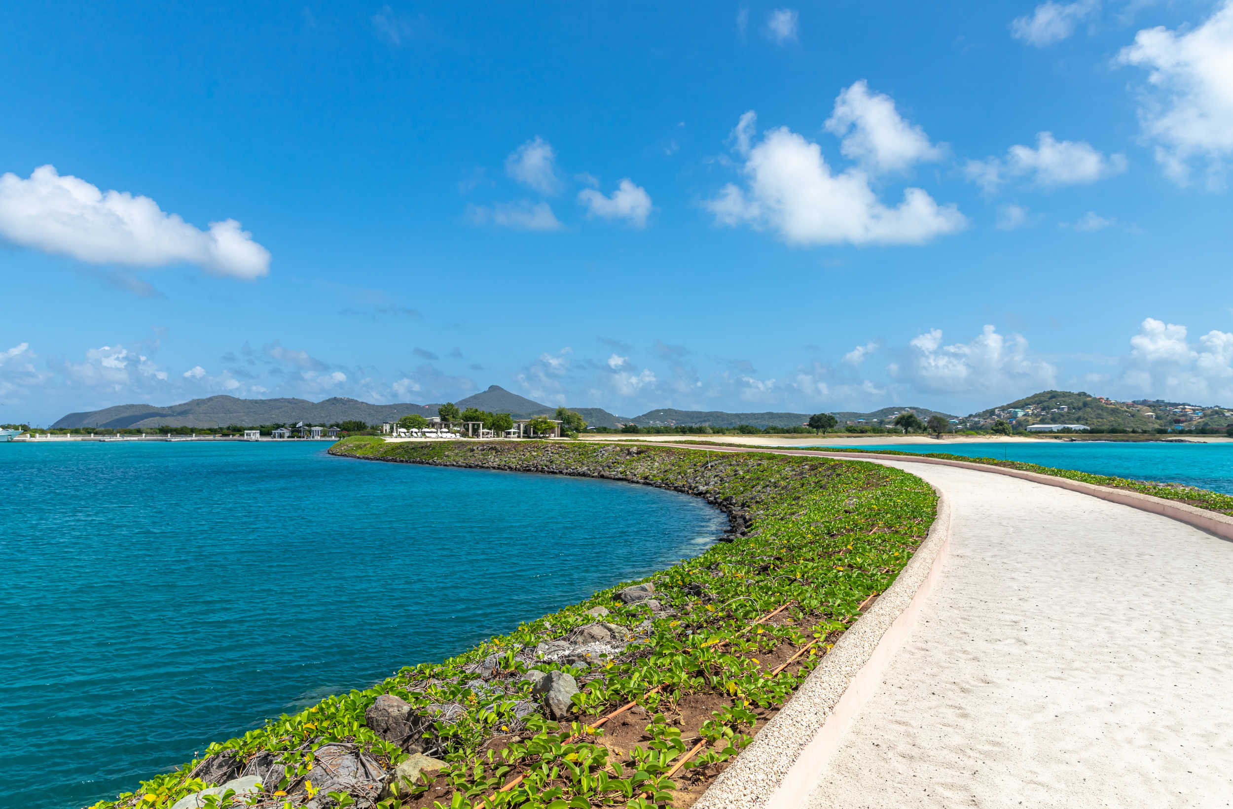 A scenic view of a paved walkway running alongside a body of turquoise water, with green plants and rocks lining the shore. In the background, there are rolling hills and mountains under a blue sky with scattered white clouds.
