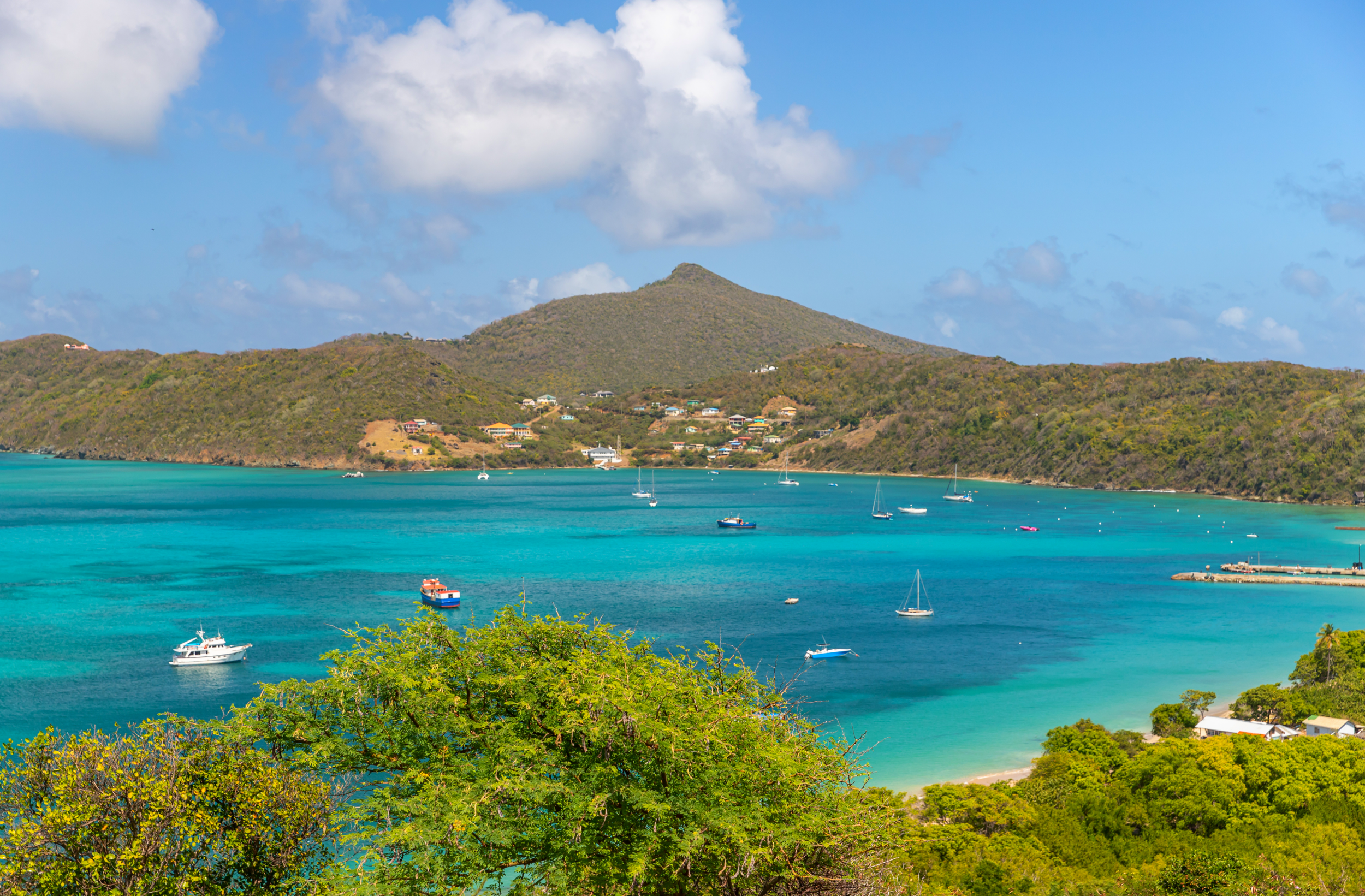 A scenic view of a turquoise bay with sailboats and yachts, surrounded by green hills and a mountain, under partly cloudy blue sky.