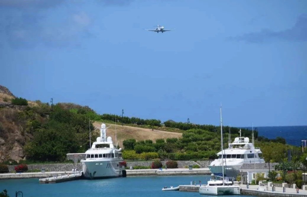 A small airplane flying in the sky above a marina with two large yachts docked. The marina is surrounded by green hills and a blue ocean, under a partly cloudy sky.