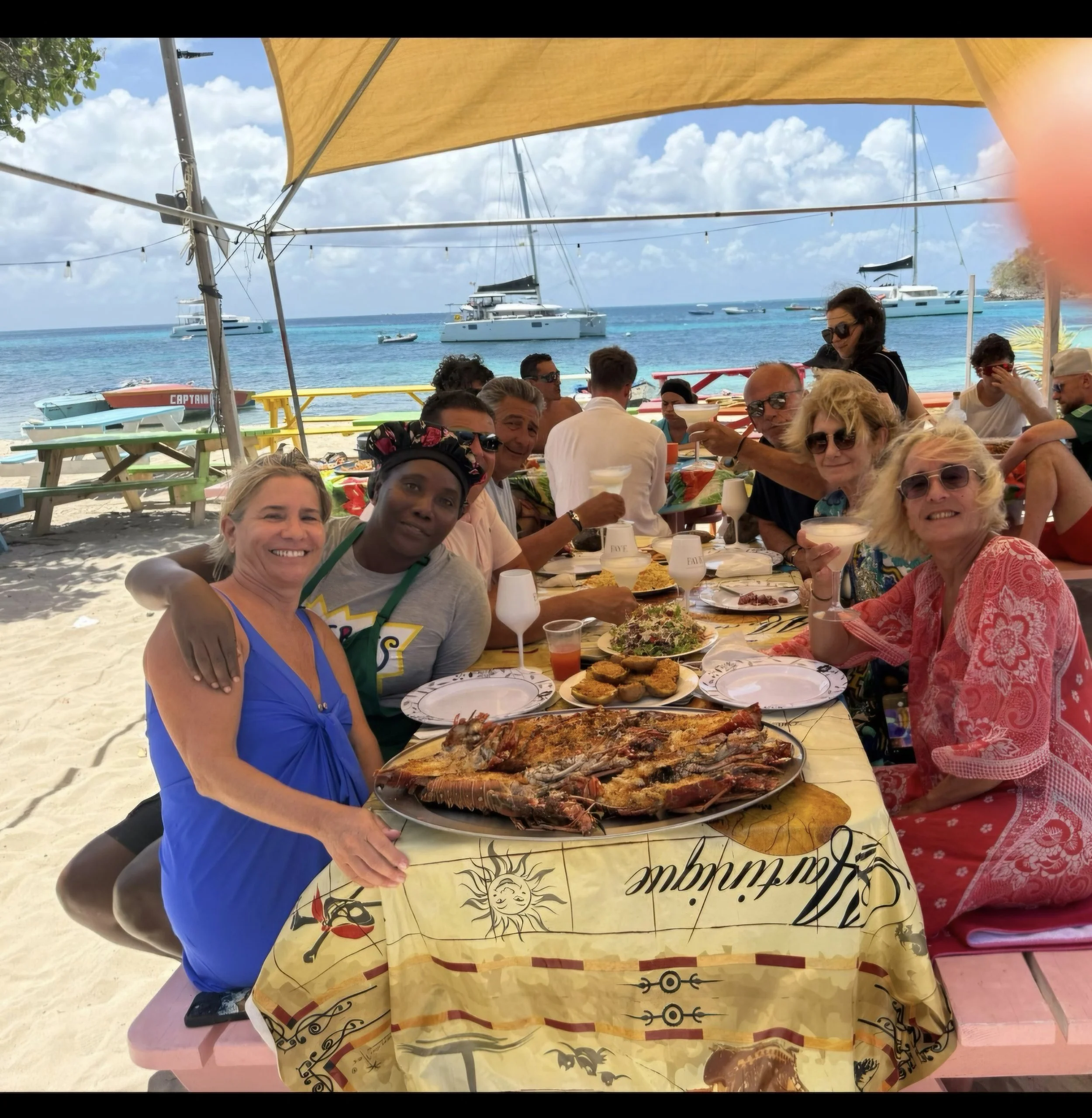 Group of people sitting at table with seafood and drinks at beachside restaurant, boats anchored in clear blue water, partly shaded by umbrella, sunny sky.