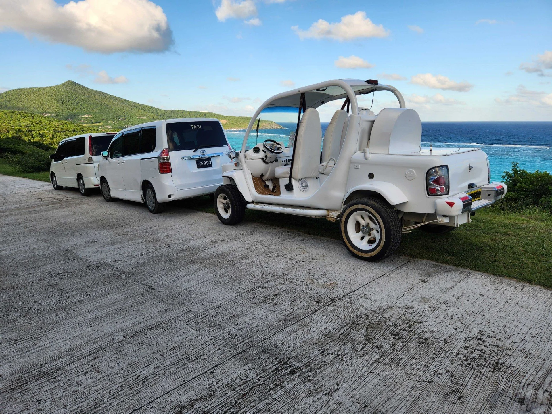 A scenic coastal view with two white taxis and a white golf cart parked on a concrete road, overlooking the ocean with green hills and a partly cloudy sky in the background.