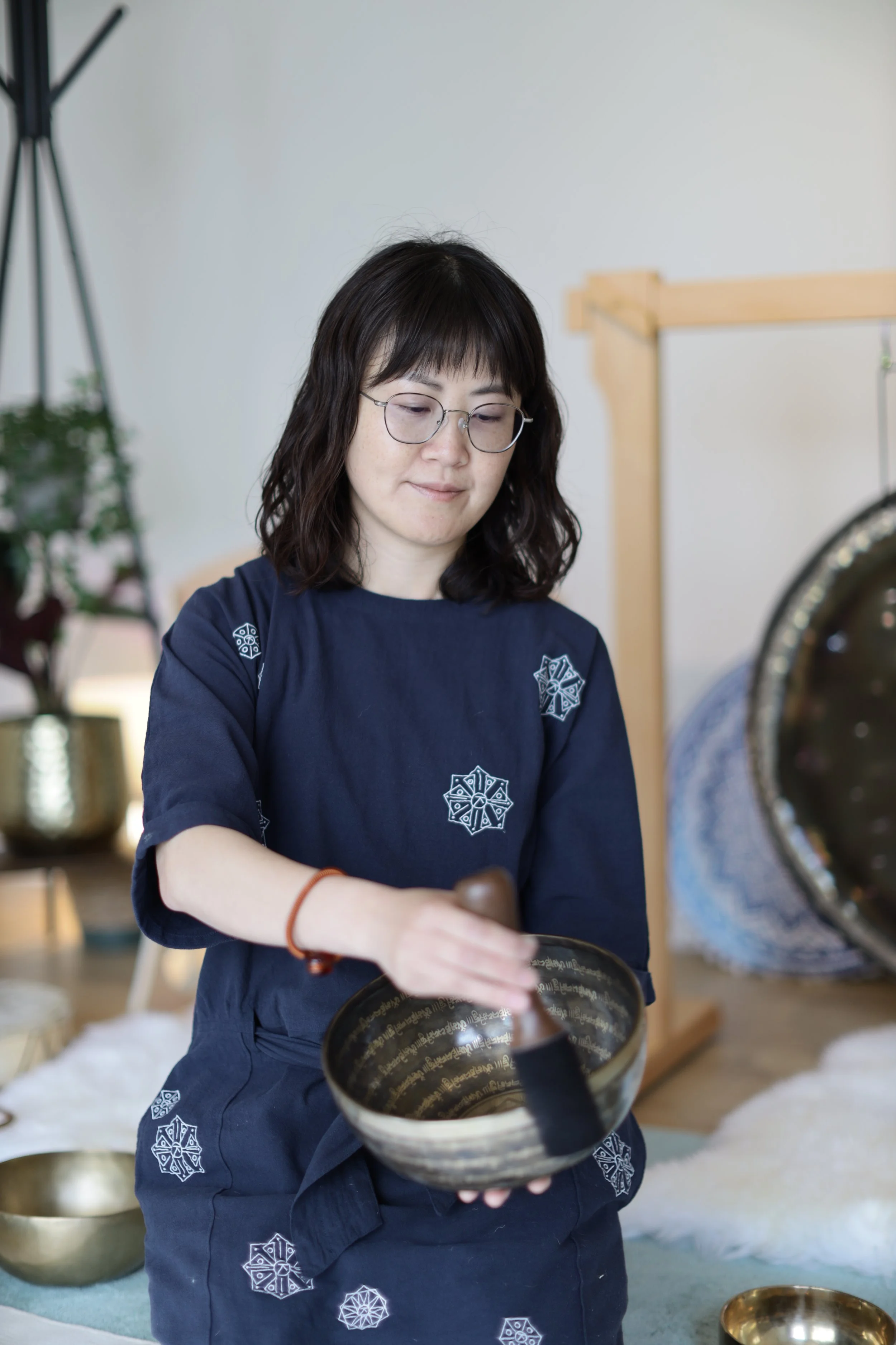 A woman with dark hair and glasses wearing a navy blue shirt with white patterns, holding a singing bowl in a bright room decorated with plants and other singing bowls.