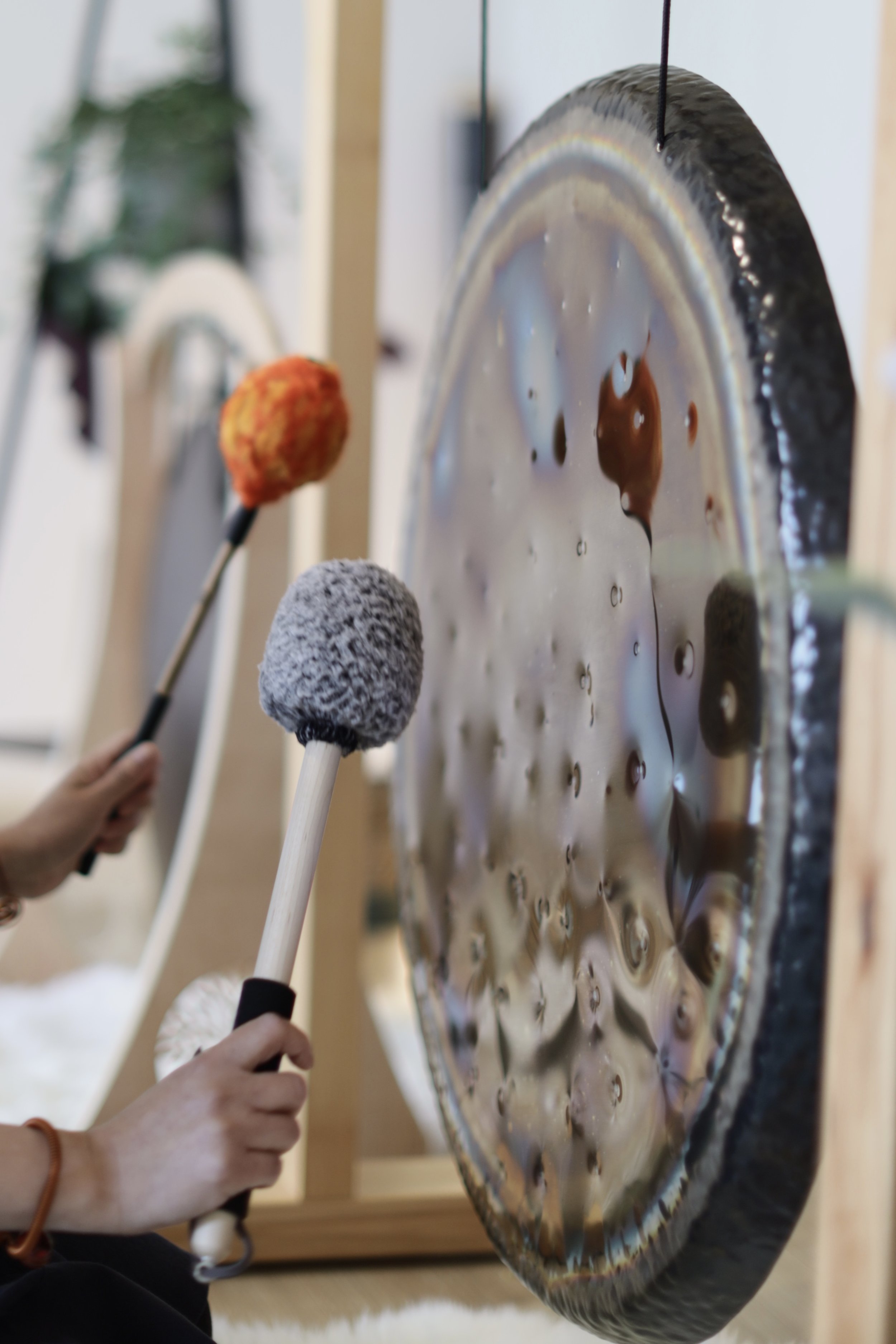 People playing a circular steel percussion instrument with mallets, with one mallet having an orange tuft and another a gray tuft, in a well-lit room.
