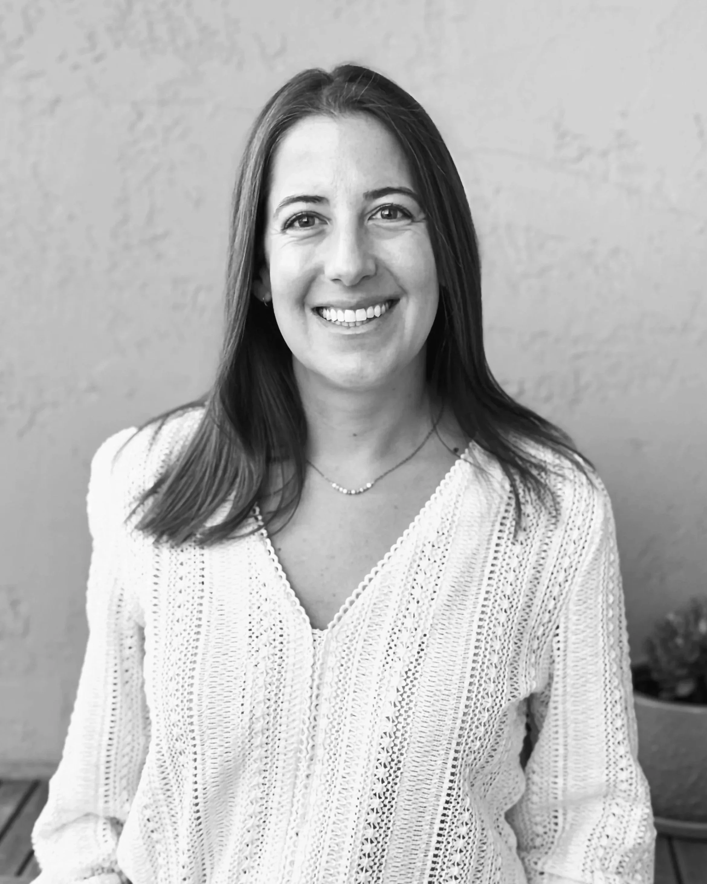 Black and white photo of a woman with long dark hair, smiling, wearing a light-colored crocheted sweater, standing against a plain wall.