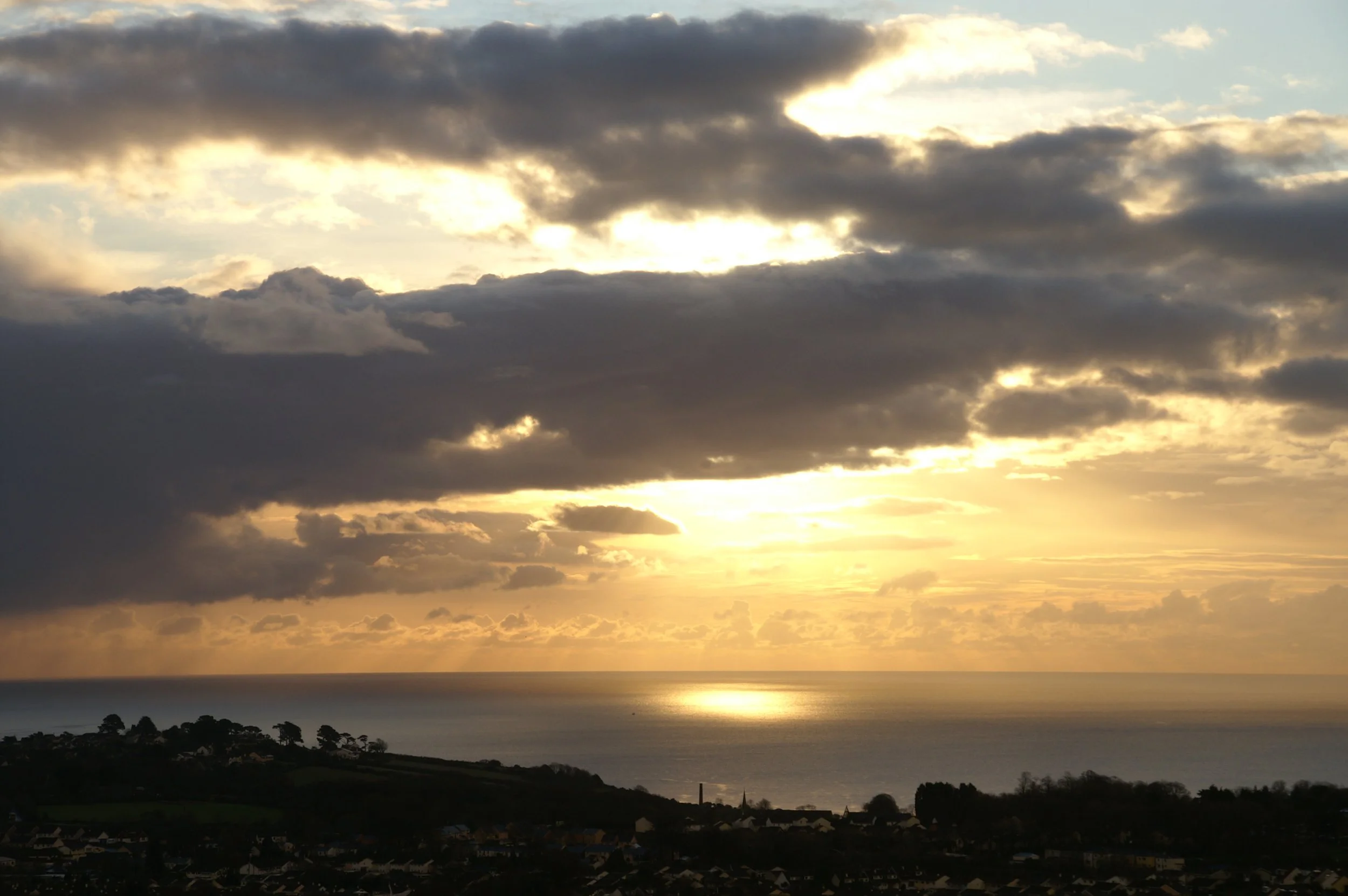 A scenic view of a sunset over a body of water, with a cloudy sky and silhouetted land and trees in the foreground.