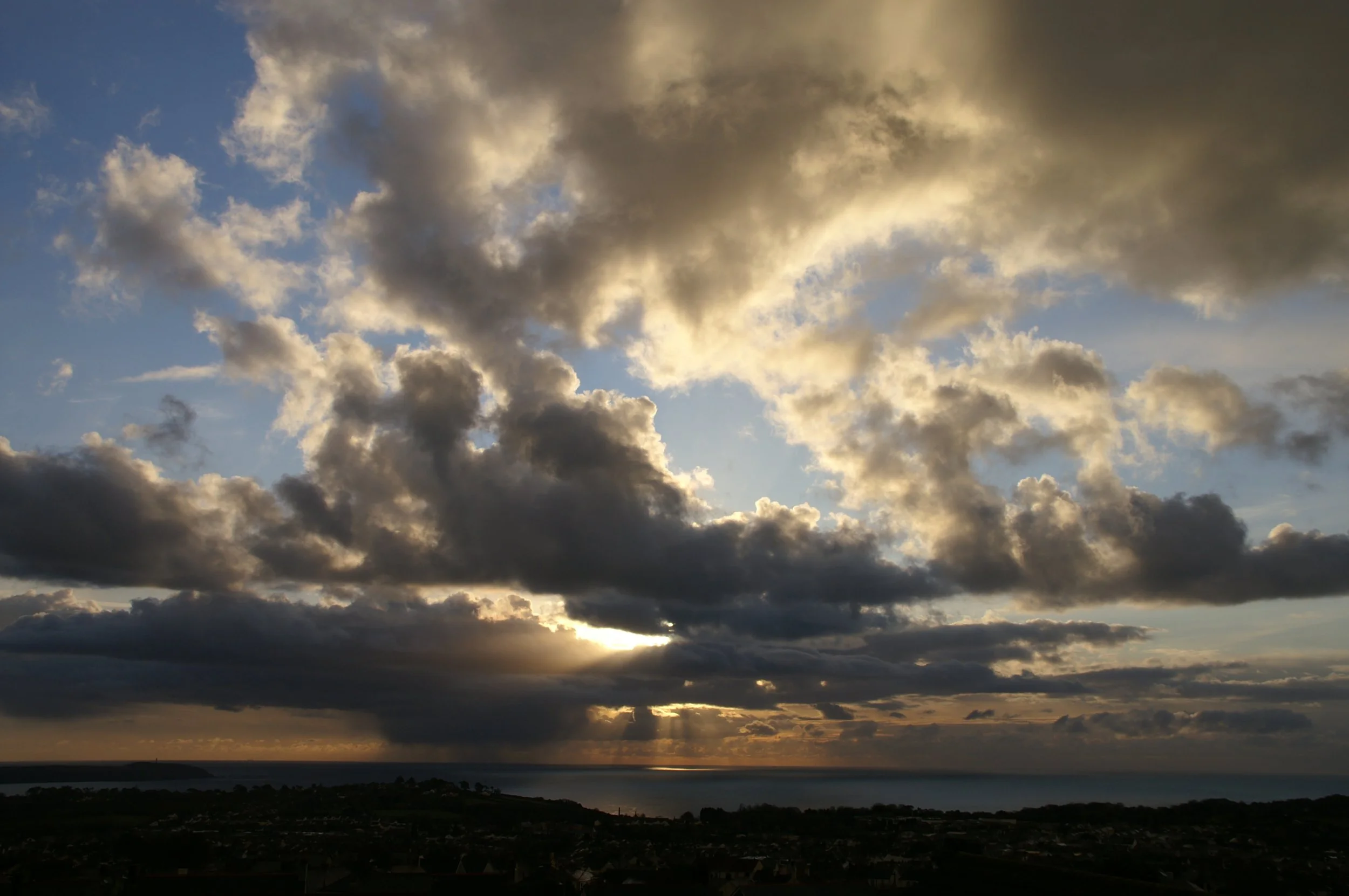 A cloudy sky during sunset with rays of sunlight peeking through the gaps in the clouds over a landscape with water and land.