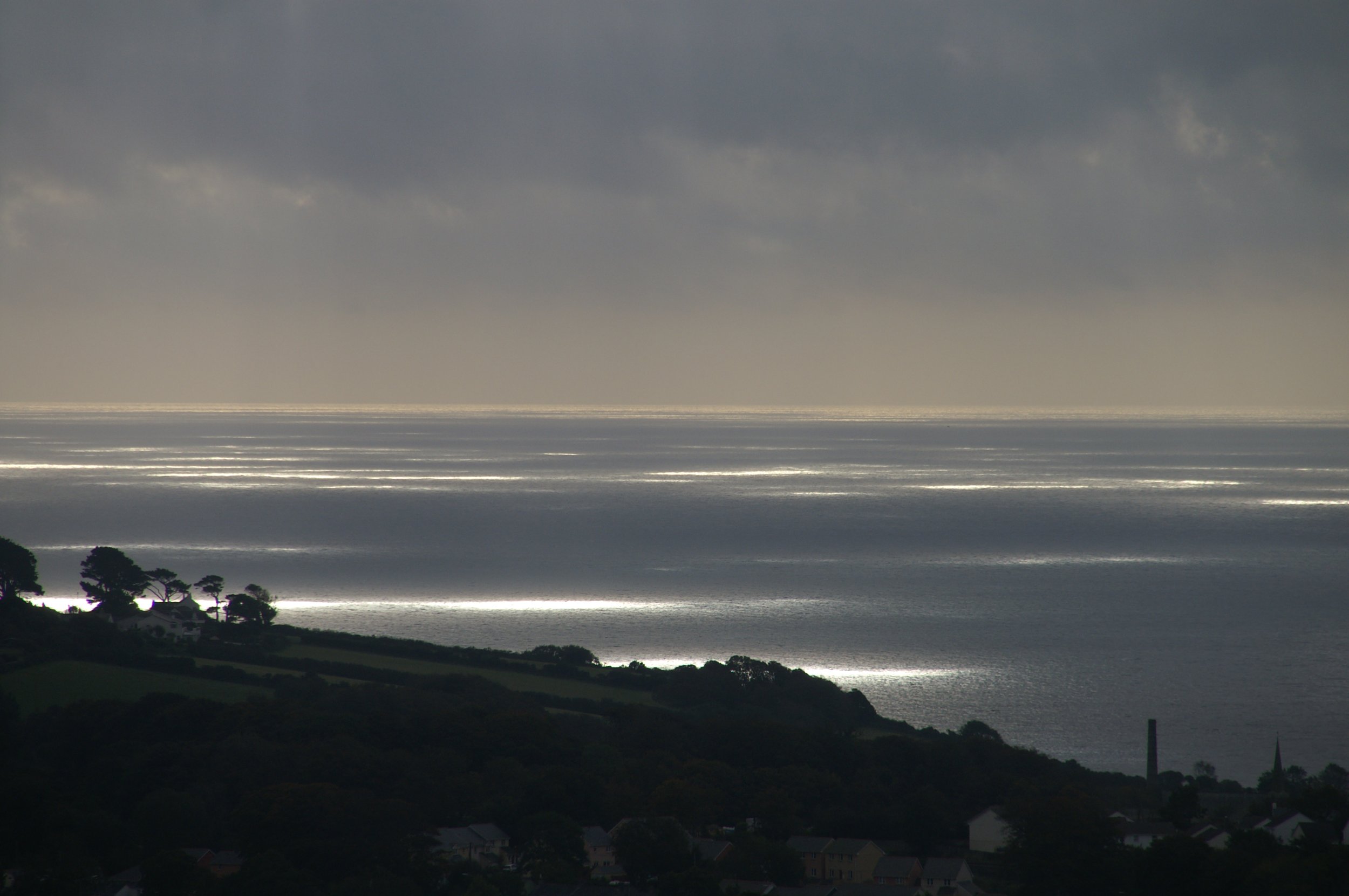 Cloudy sky over the ocean with sunlight reflecting on the water and silhouetted trees and houses in the foreground.