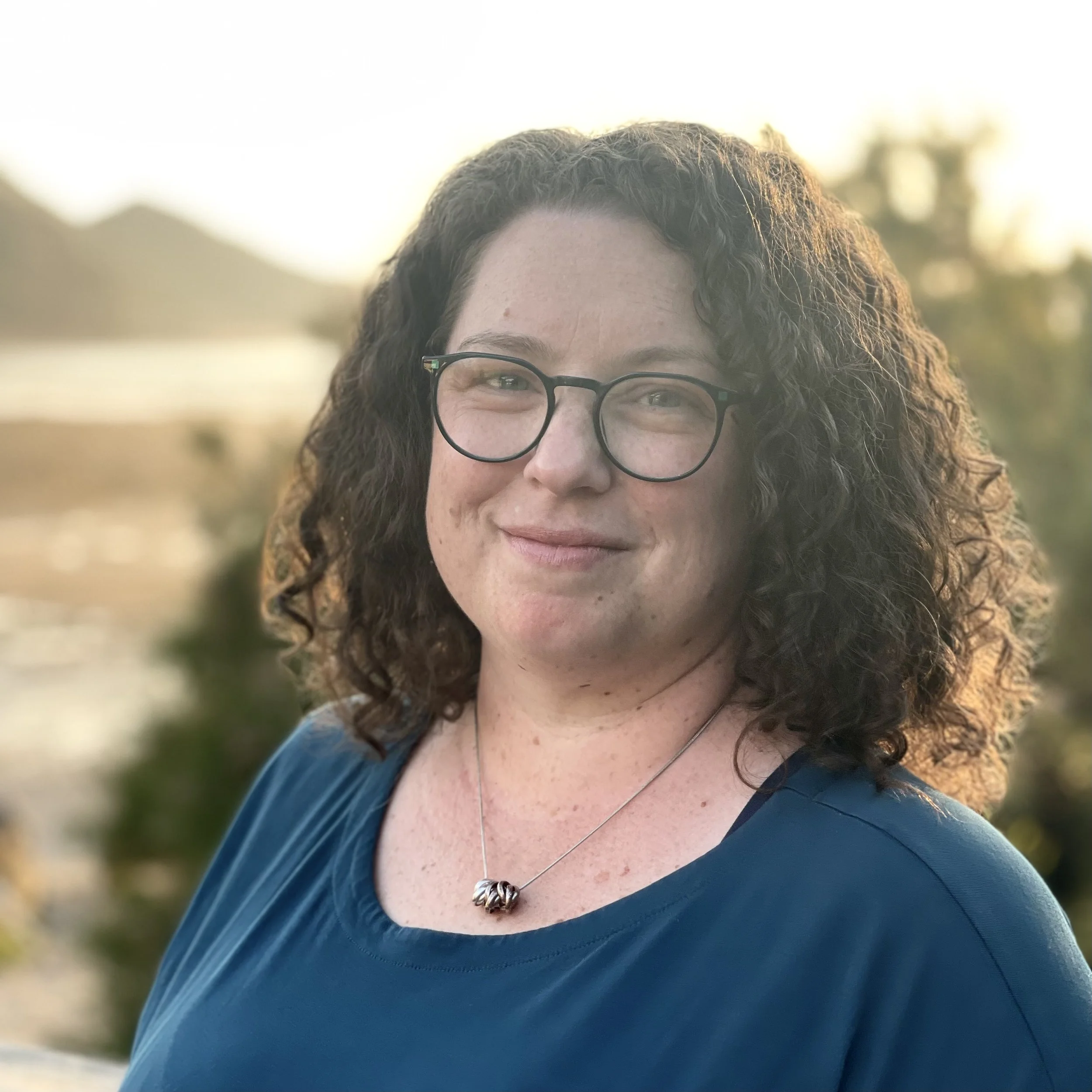 A woman with curly brown hair wearing glasses, a blue shirt and a necklace, smiling outdoors.