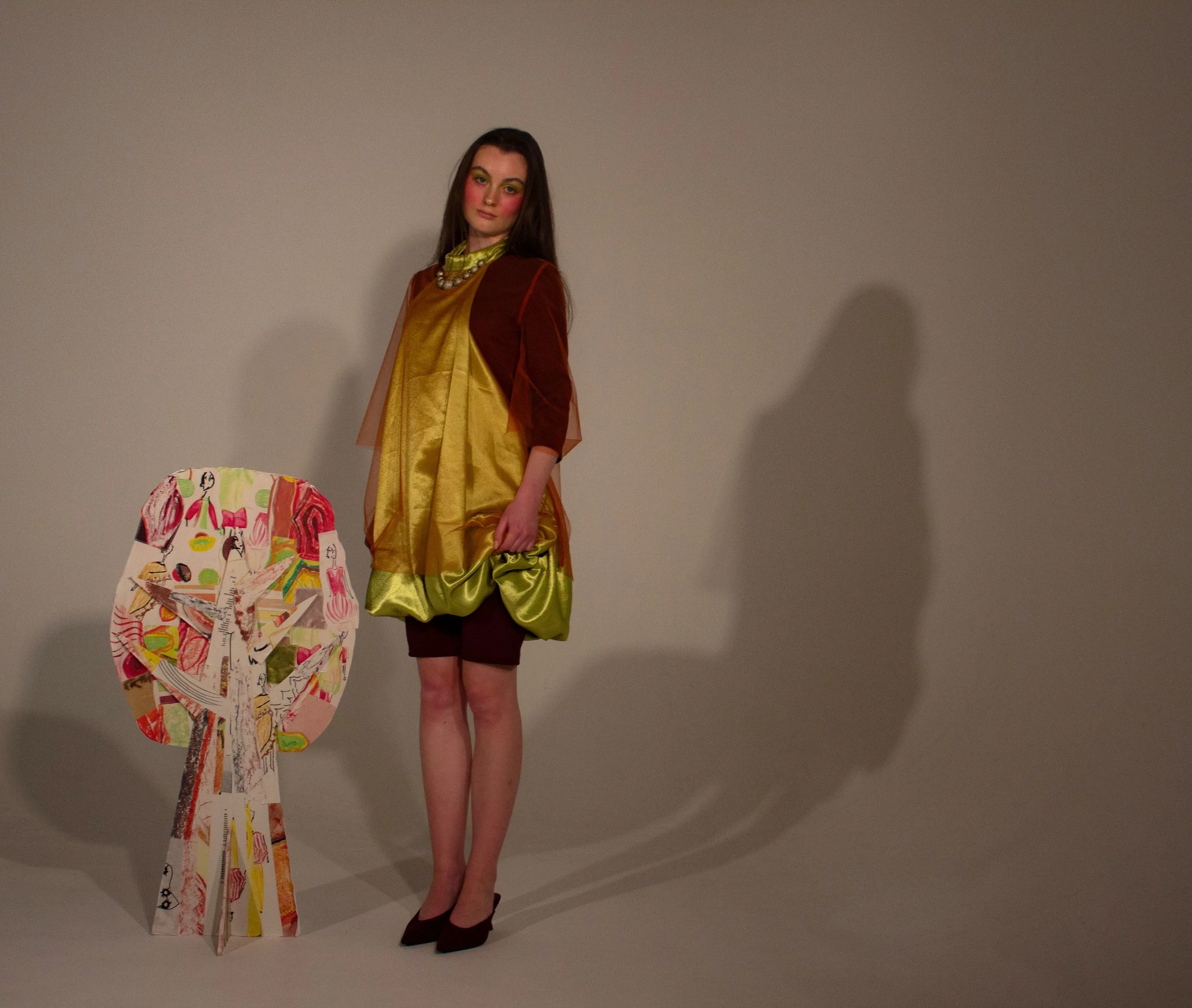 A woman with long dark hair and bright makeup stands against a plain wall, wearing a shiny green dress with a russet jumpsuit underneath, both layered with an orange tulle dress. Beside her, there is an abstract, colourful sculpture of a tree.