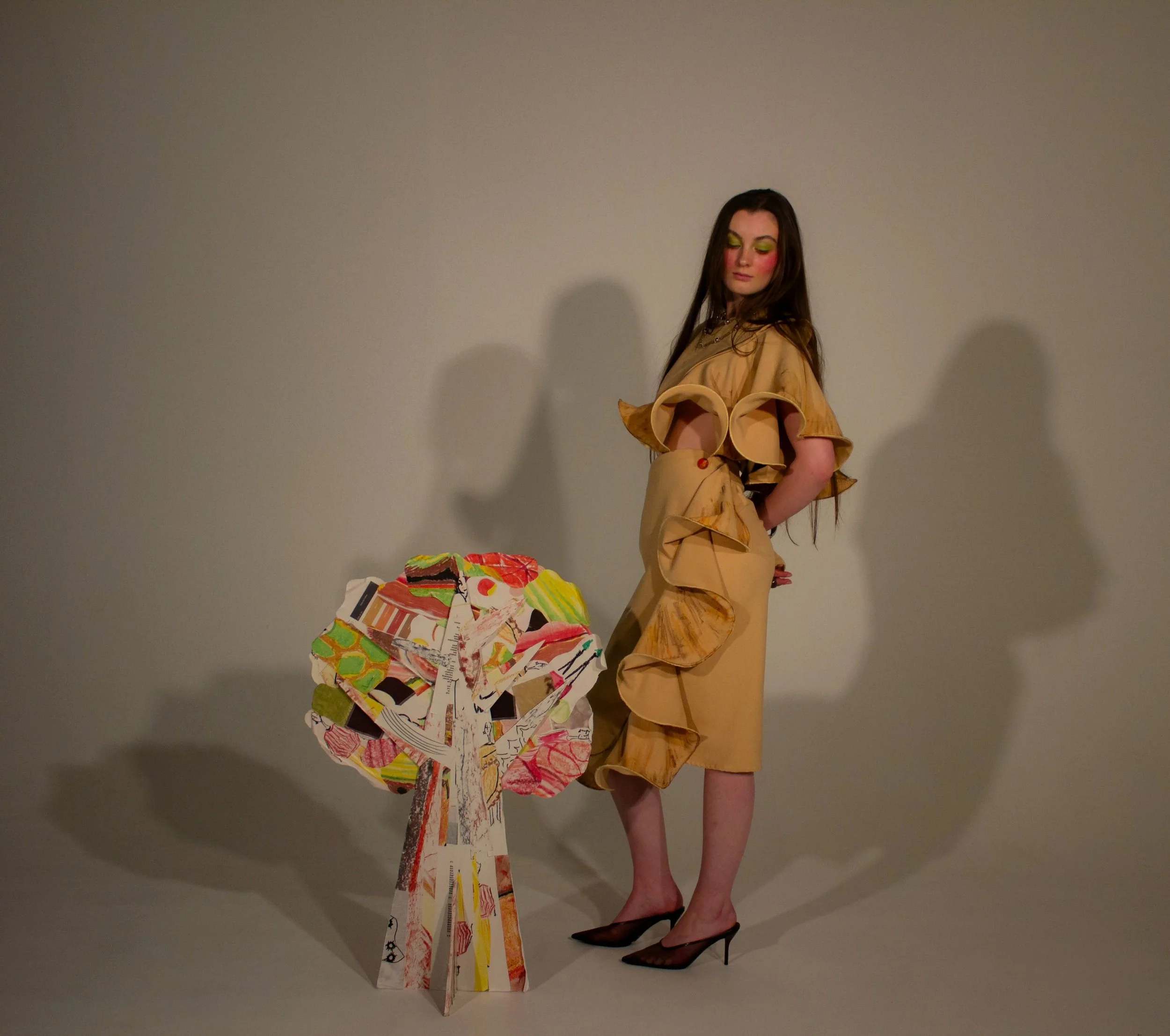 A woman with long brown hair dressed in a  beige ruffle skirt, top and one sleeved jacket, wearing brown high heels, stands next to an abstract, colorful sculpture made of various torn and painted paper pieces assembled into a tree form.