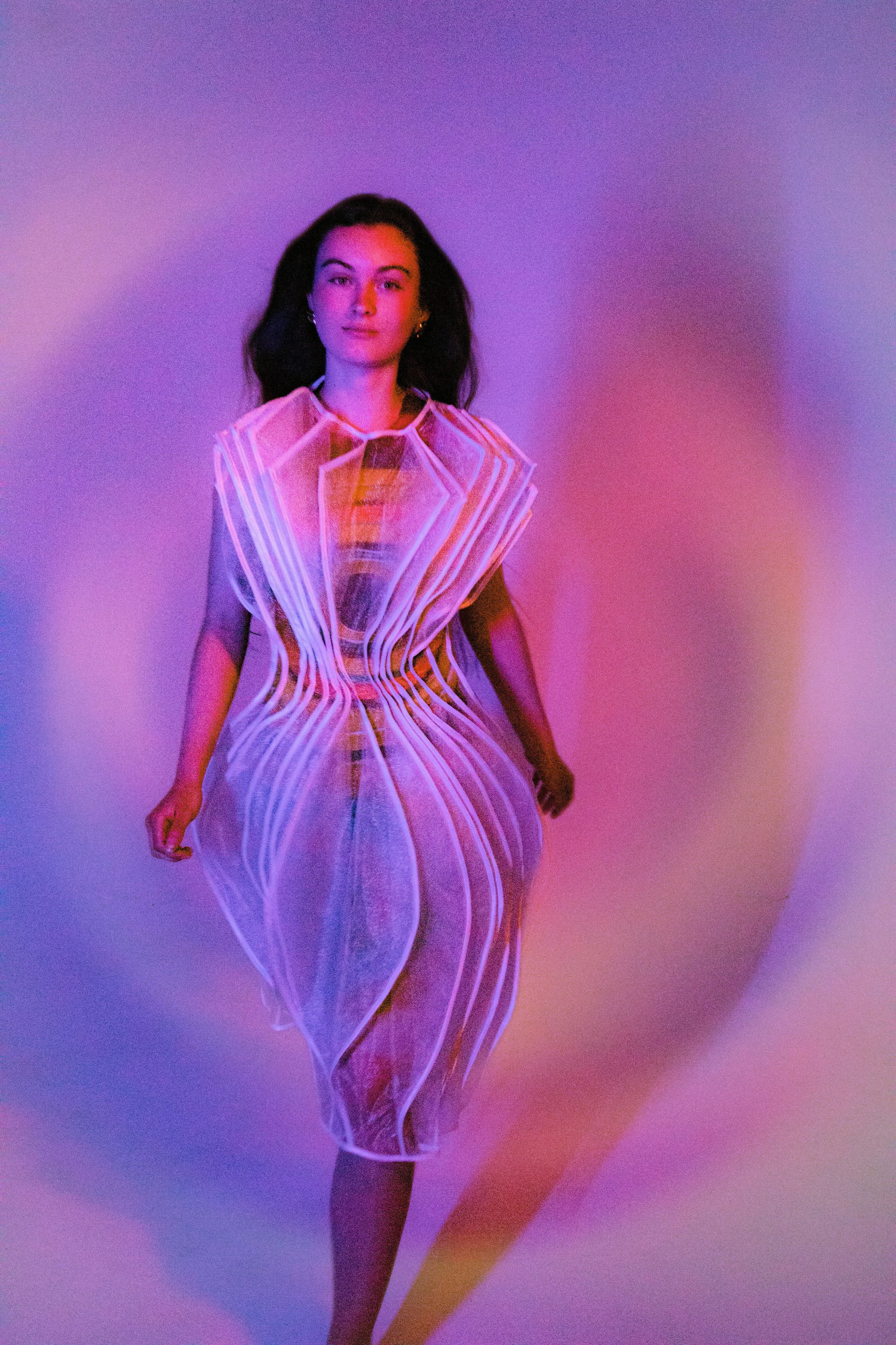 A woman in a sheer, circle shaped dress layered over an abstract circle printed dress, stands against a colourful gradient background with pink, purple, and blue hues, casting a shadow onto the wall.
