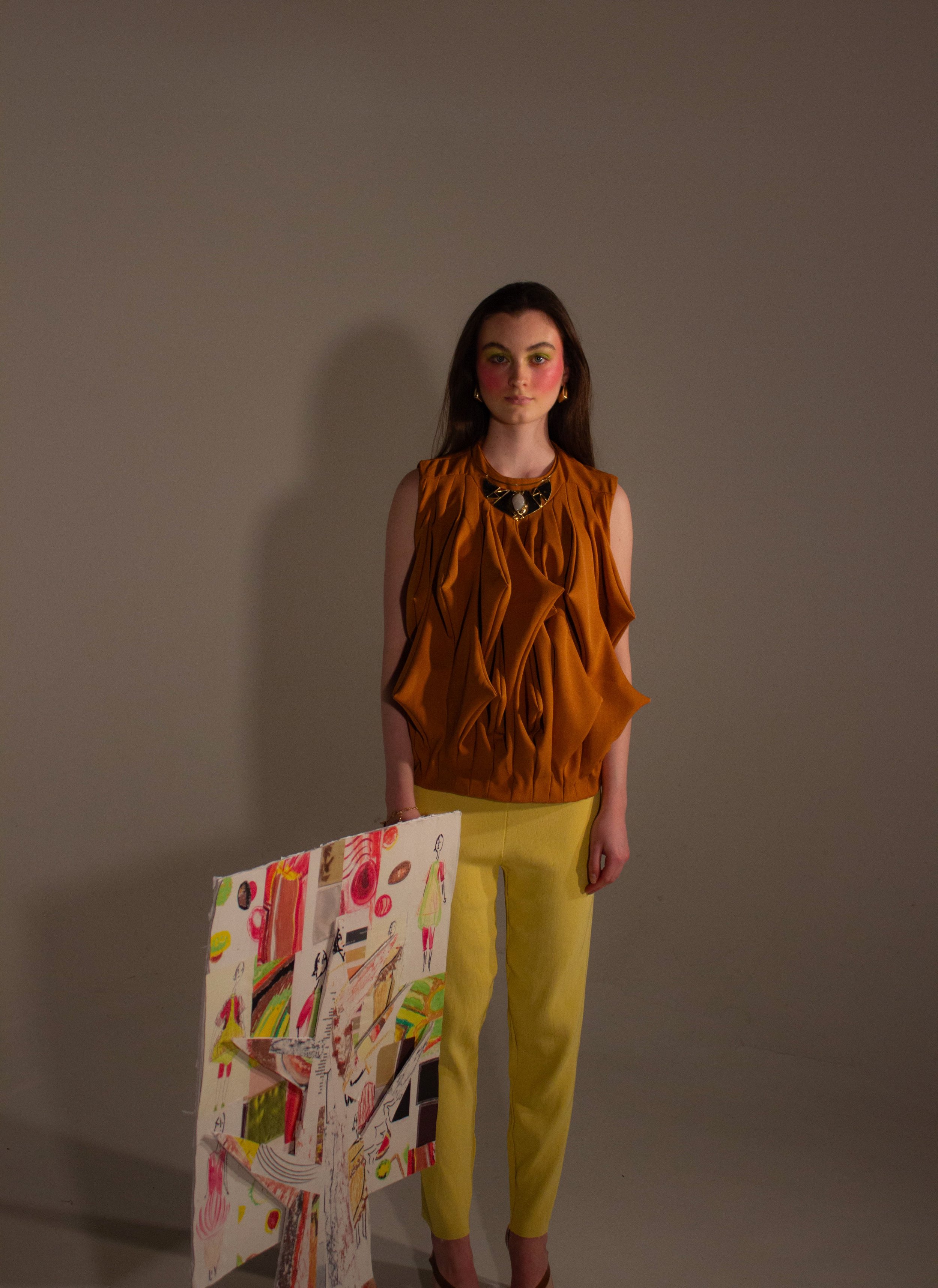 A young woman with dark brown hair wearing a sleeveless orange top made of triangle shapes, gold earrings, and yellow ribbed leggings, stands indoors against a plain wall, with colorful artwork tree on display to her left.