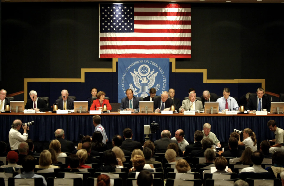 Panel discussion of the New Mexico Department of Justice with formal attendees, with the American flag and the National Commission on Terrorist Attacks banner in the background.