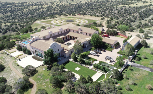 A large estate representing Zorro Ranch in Stanley, New Mexico with multiple buildings, parking lot, surrounding greenery, and a circular landscaped area in the distance.