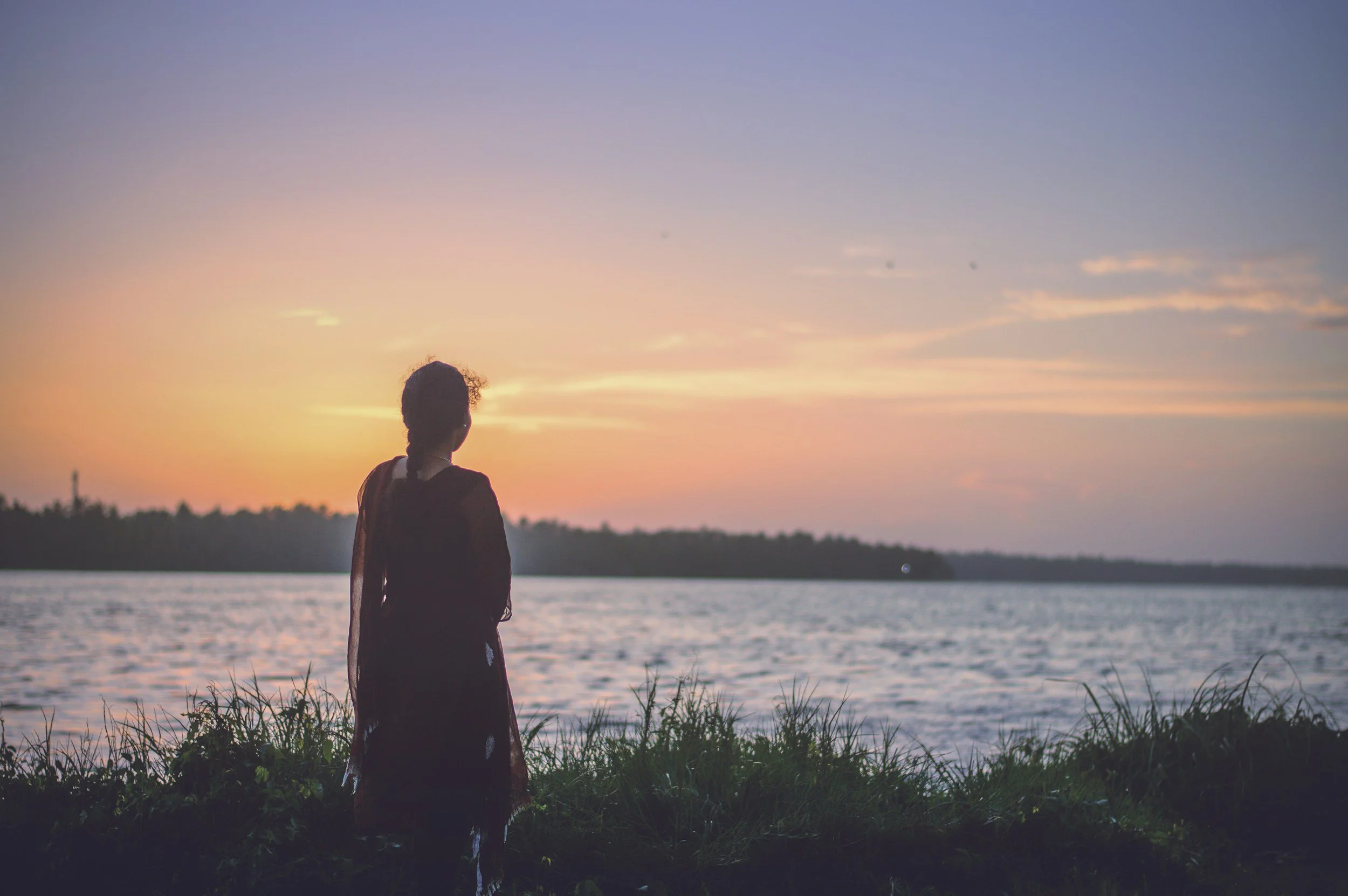Woman standing by calm water at sunset, representing reflection, emotional healing, and inner calm.
